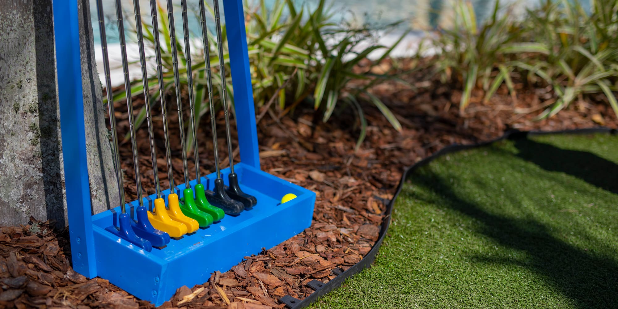 Blue rack with colorful mini golf putters on bark mulch near a putting green.