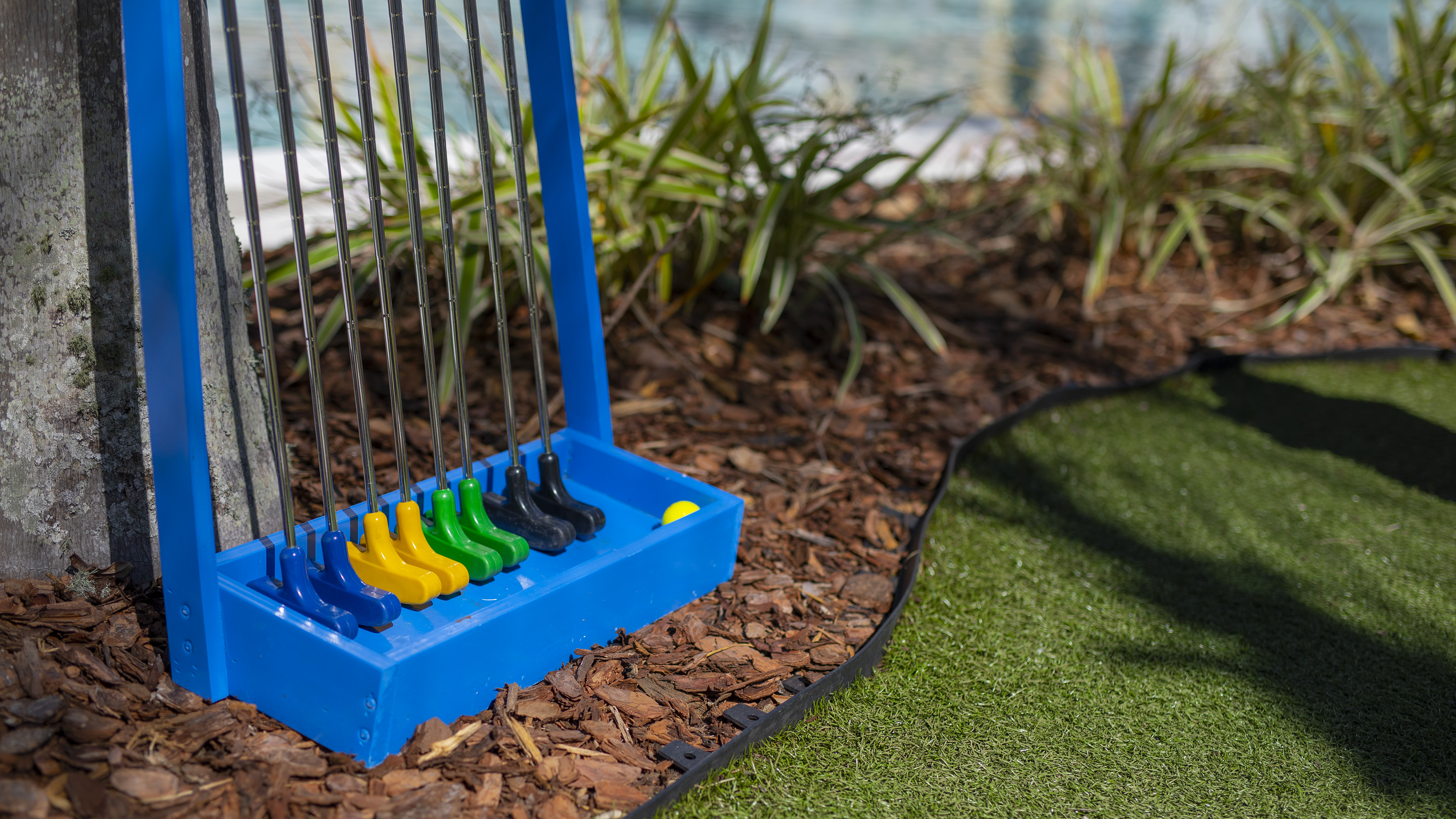 Blue rack with colorful mini golf putters on bark mulch near a putting green.