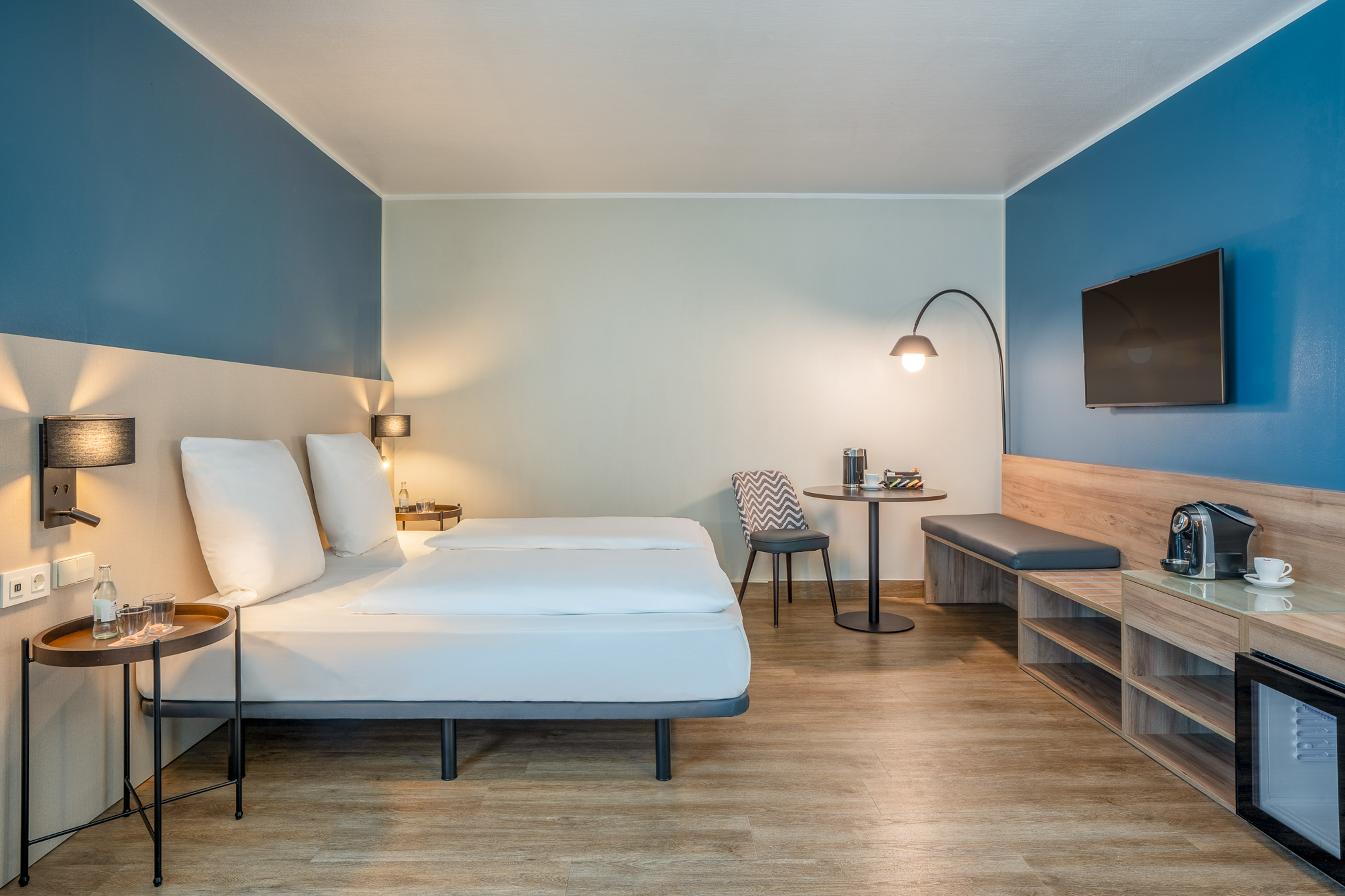 Modern hotel room with twin beds, blue wall, TV, mini-fridge, and coffee machine.