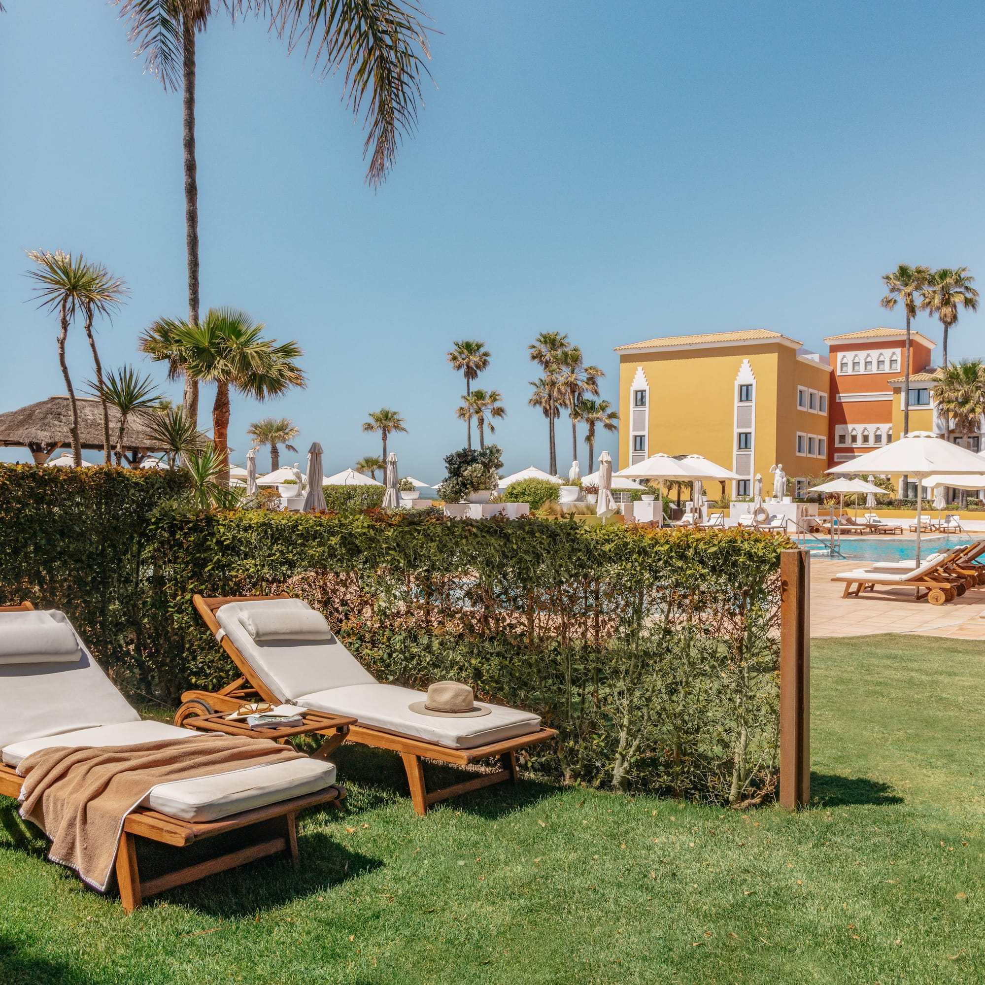 Tropical resort: sun loungers on grass, hotel pool, palm trees, blue sky.