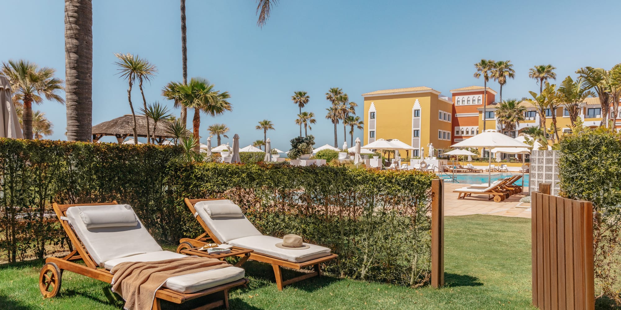Tropical resort: sun loungers on grass, hotel pool, palm trees, blue sky.