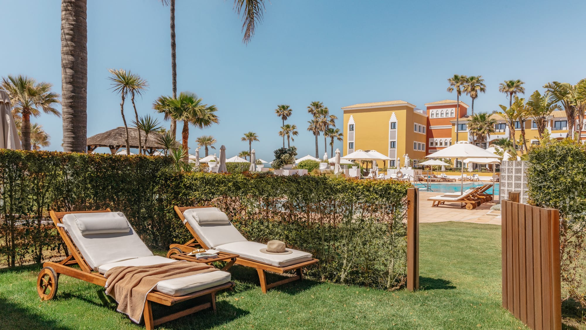 Tropical resort: sun loungers on grass, hotel pool, palm trees, blue sky.