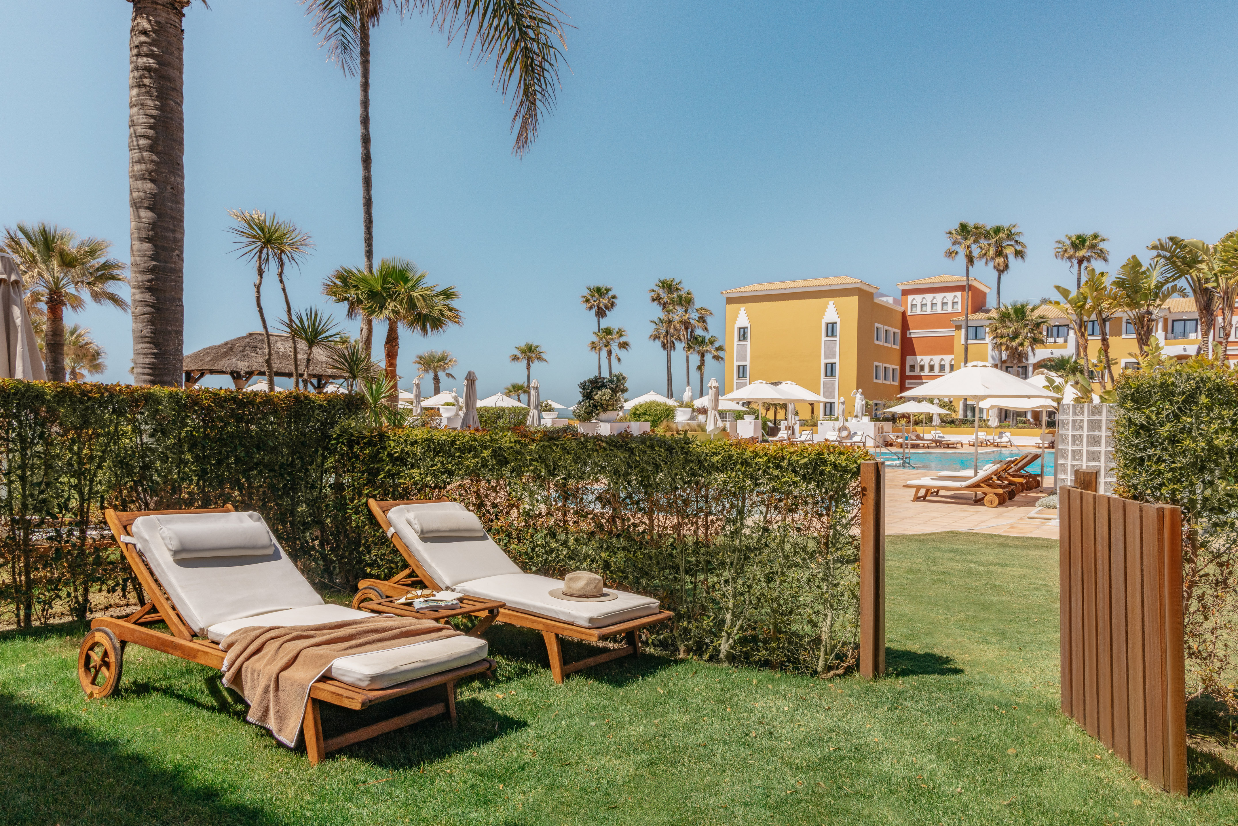 Tropical resort: sun loungers on grass, hotel pool, palm trees, blue sky.