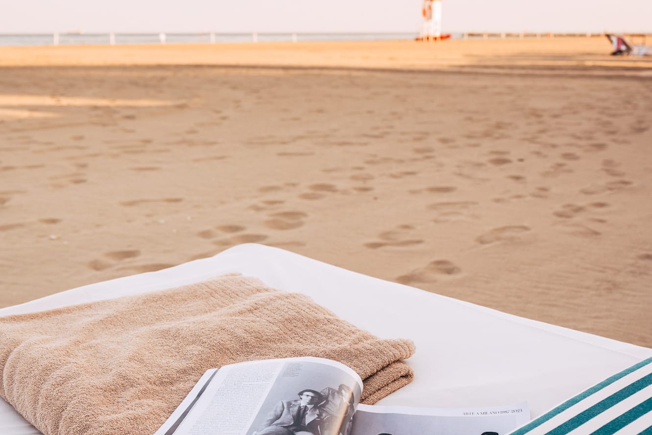Beach bed with open magazine, brown towel, and striped pillow on sandy shore.