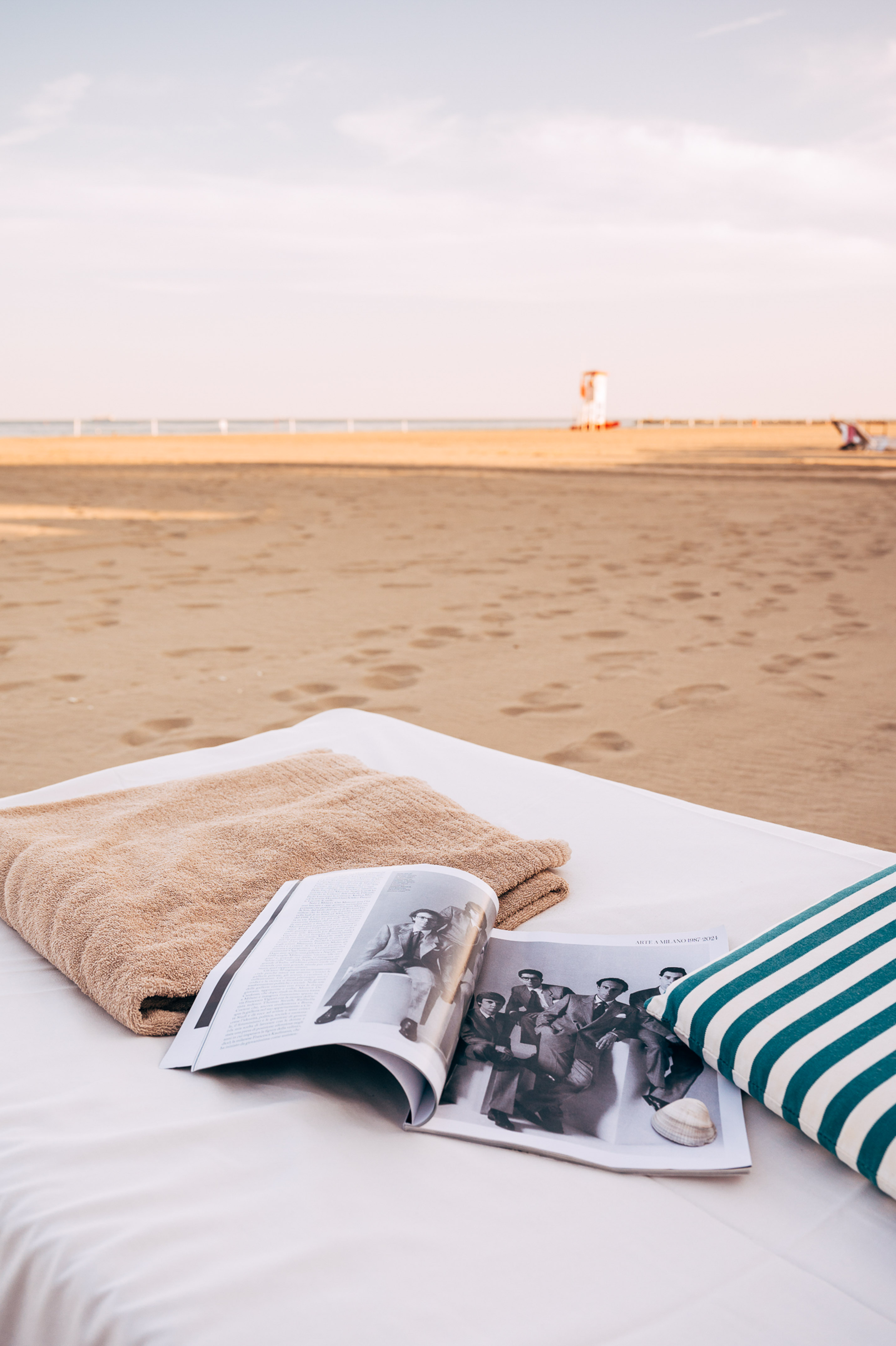 Beach bed with open magazine, brown towel, and striped pillow on sandy shore.