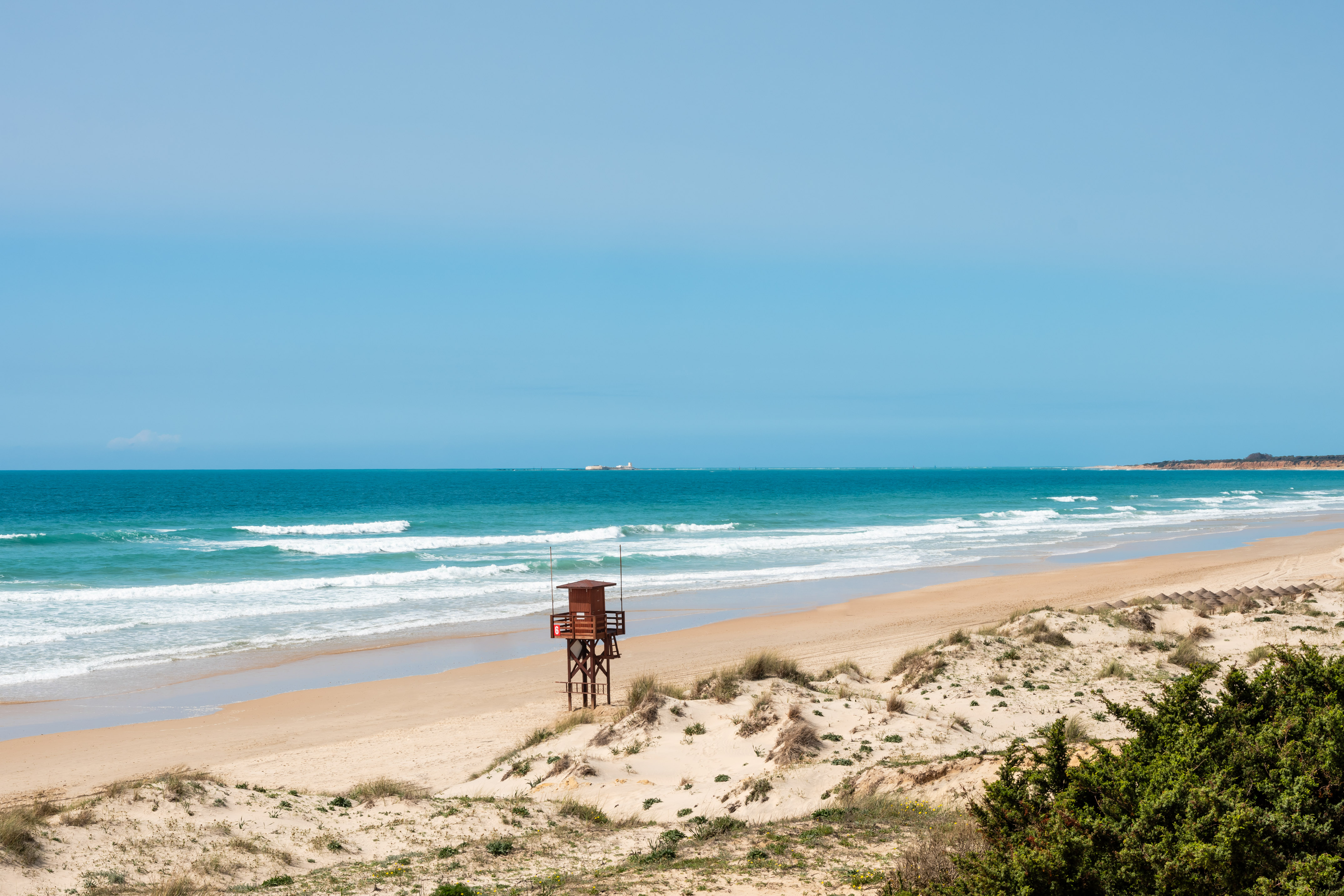 Sandy beach with a wooden lifeguard tower, blue ocean, and grassy dunes.