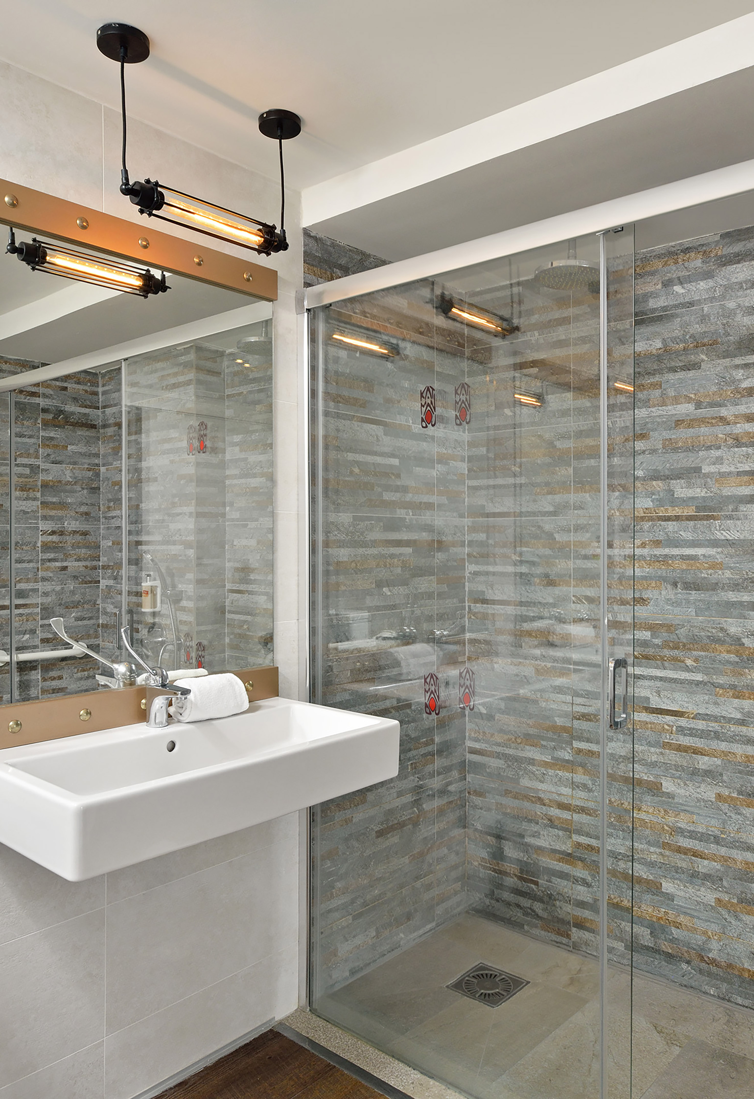 Modern bathroom: white sink, industrial lights, glass shower, stone tiles.