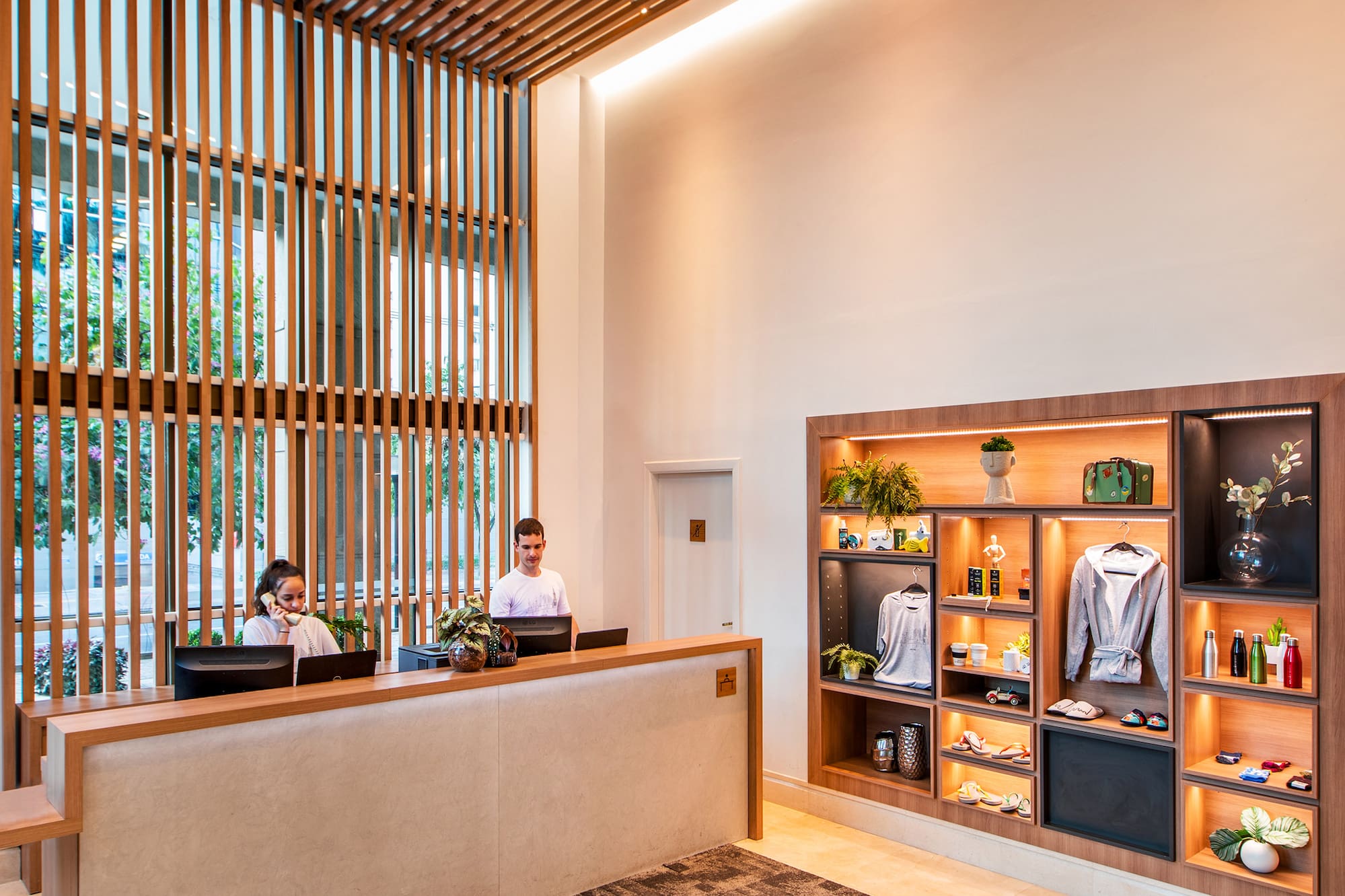 Modern hotel reception desk, two staff, wood slat ceiling, and display shelves.