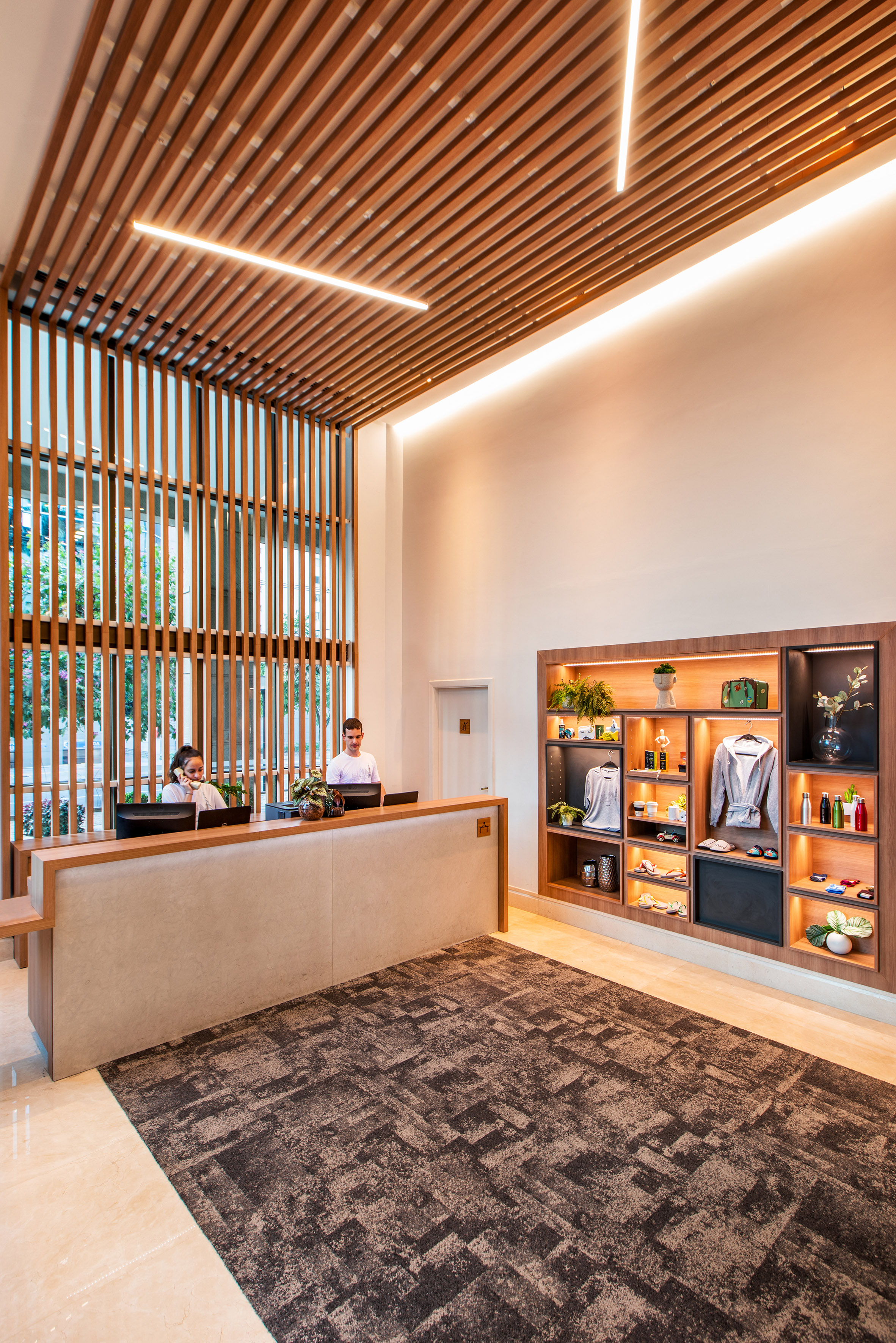 Modern hotel reception desk, two staff, wood slat ceiling, and display shelves.