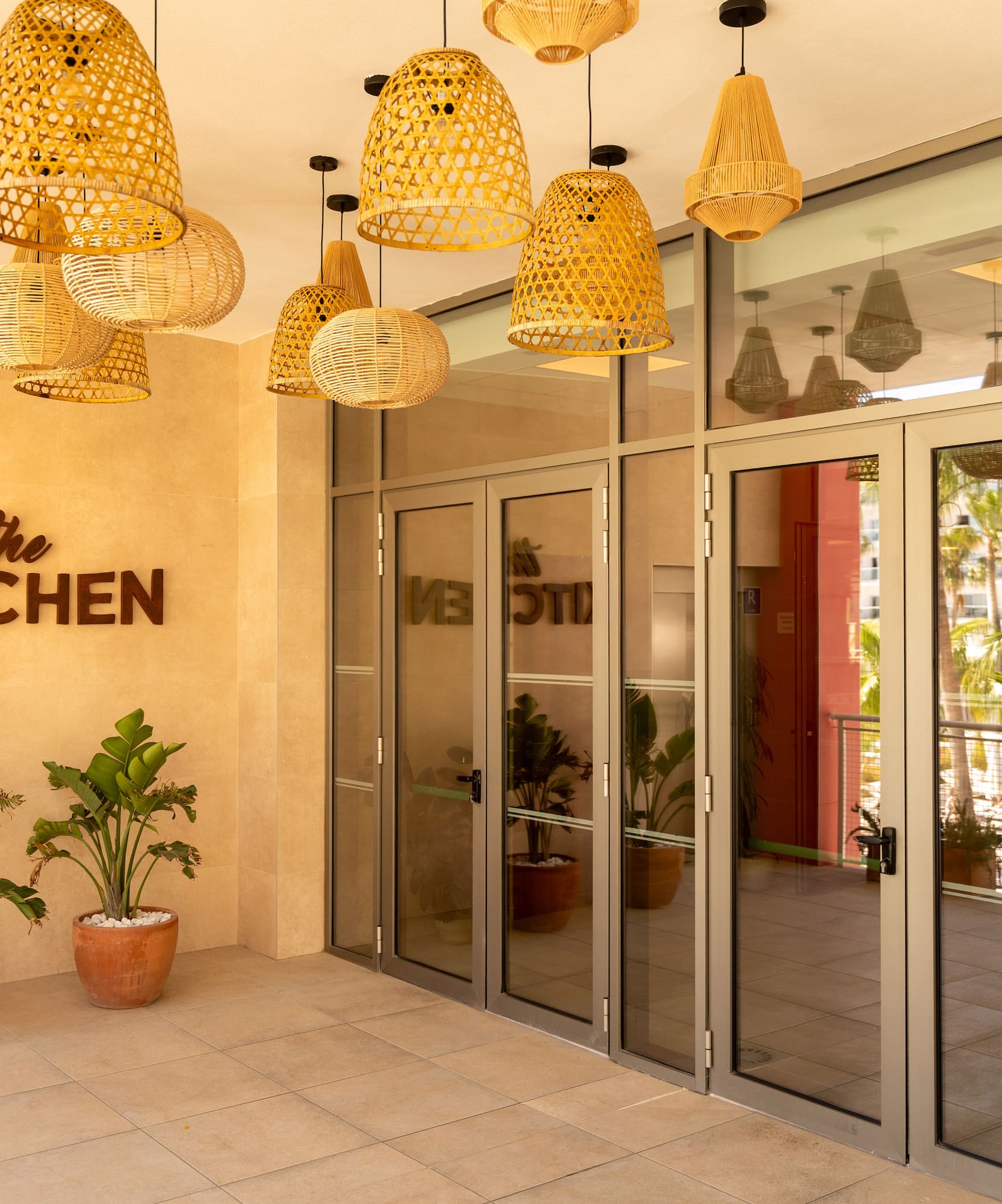 Restaurant 'The Kitchen' entrance, wicker pendant lights, tropical plants, and pool.