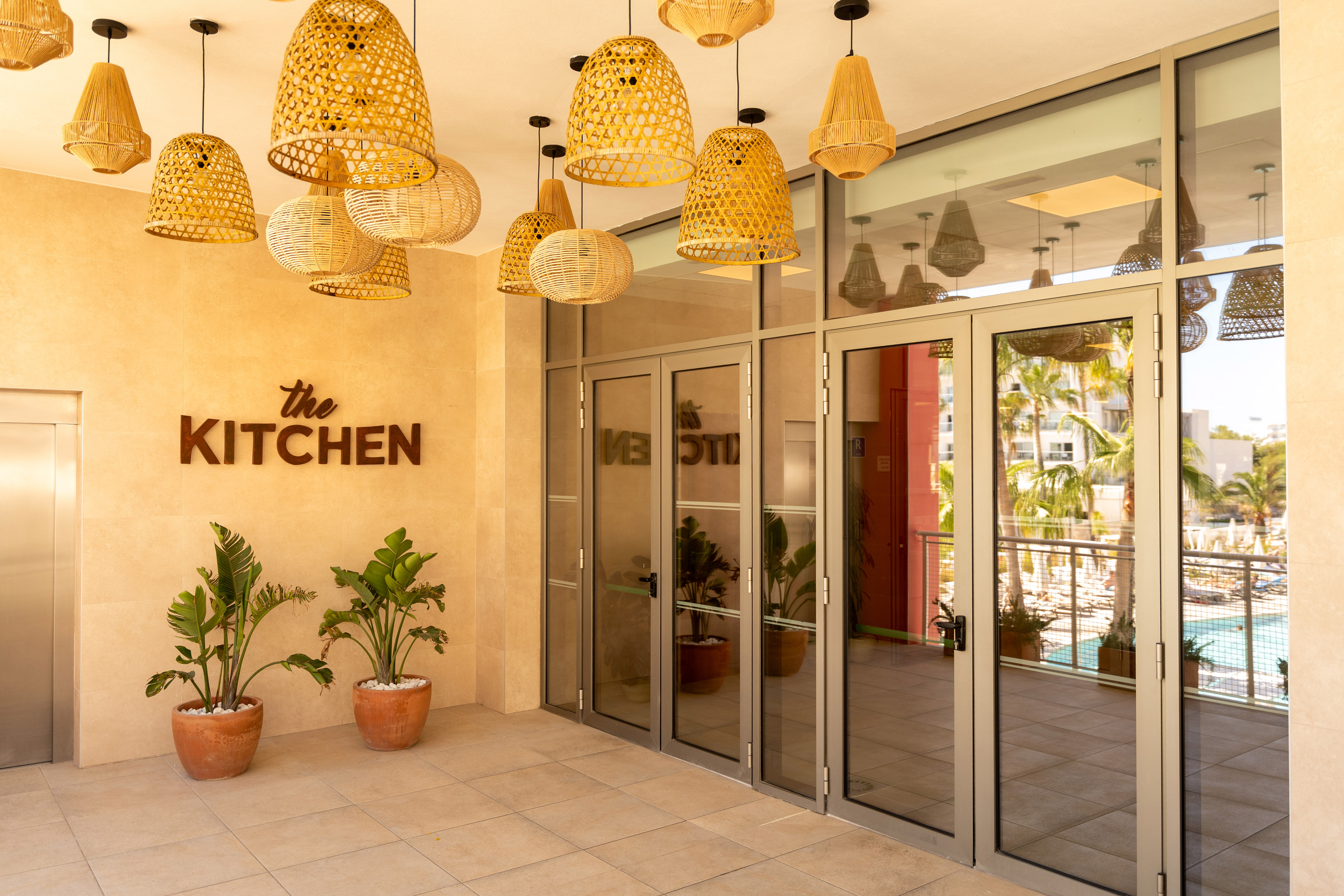 Restaurant 'The Kitchen' entrance, wicker pendant lights, tropical plants, and pool.