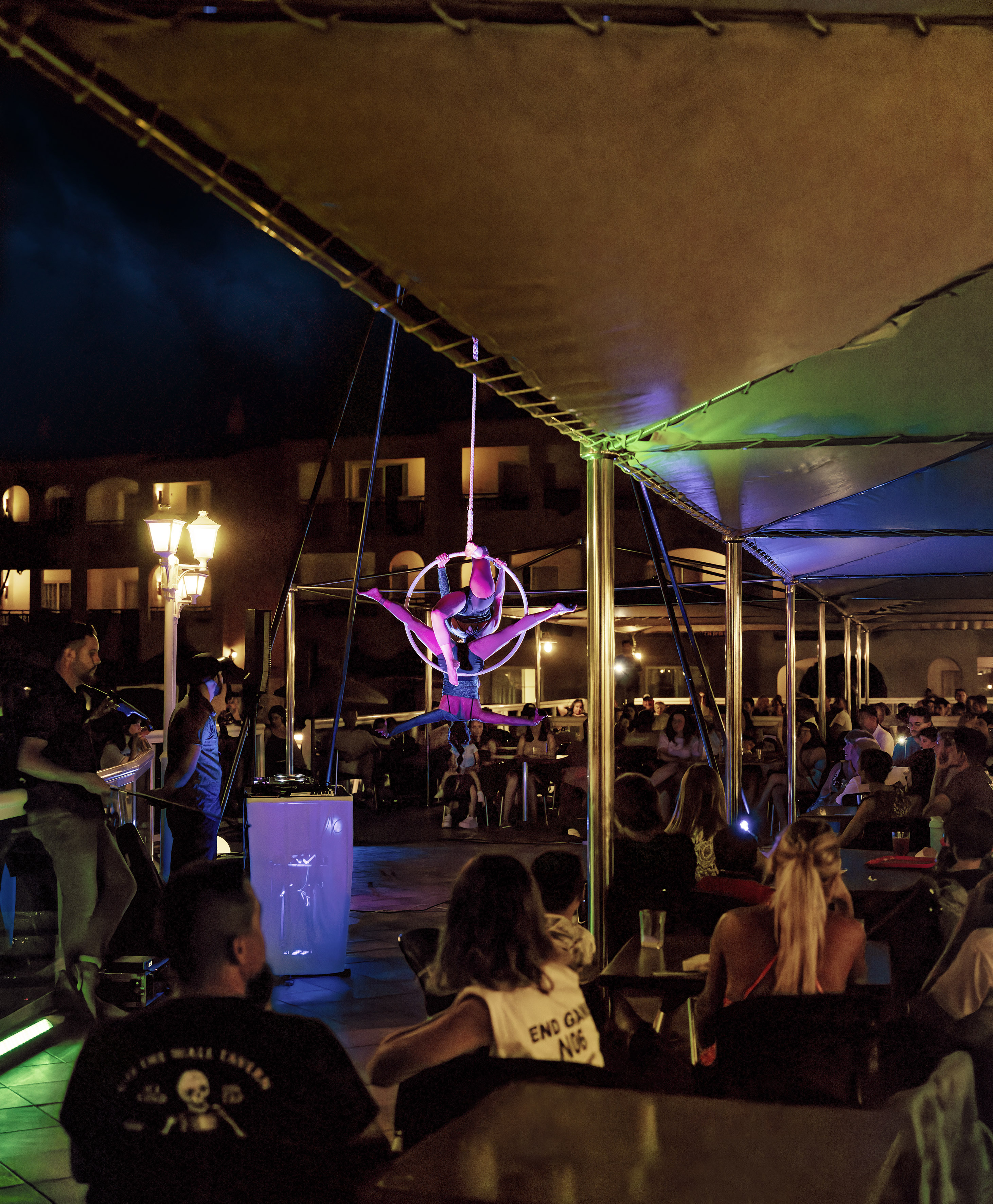 Aerial hoop performer in pink light, entertaining an outdoor night audience.