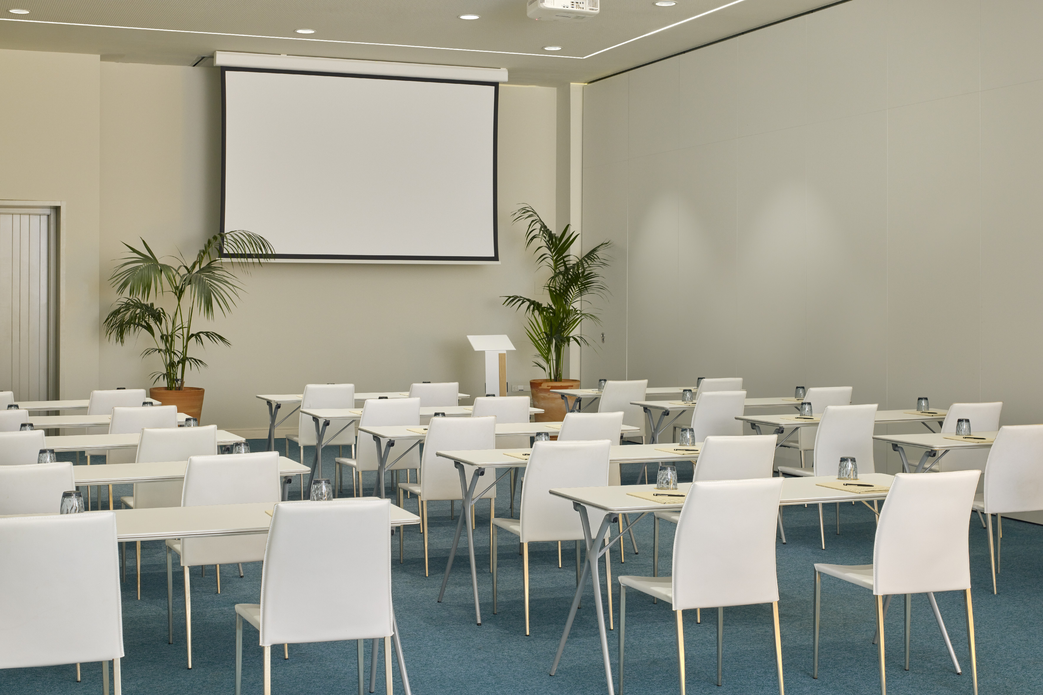 Modern conference room with screen, white tables, chairs, blue carpet.