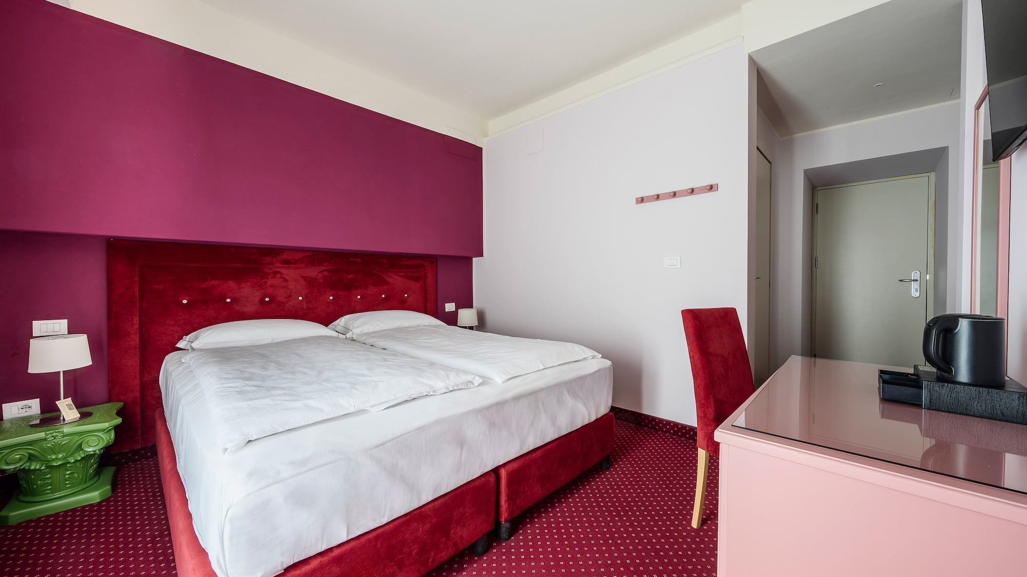 Hotel bedroom with red velvet double bed, magenta wall, and patterned carpet.