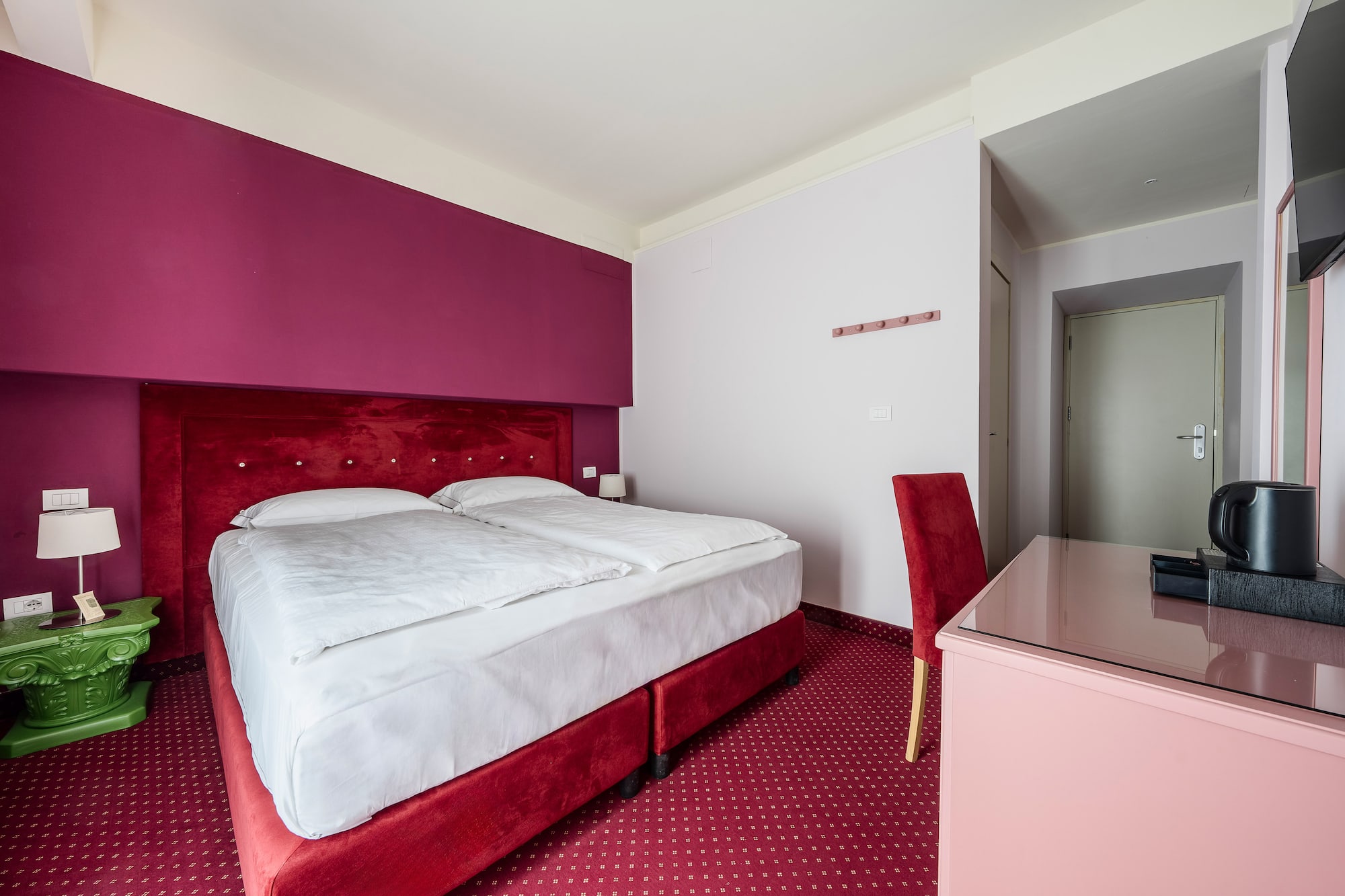Hotel room with red velvet bed, magenta wall, and patterned red carpet.