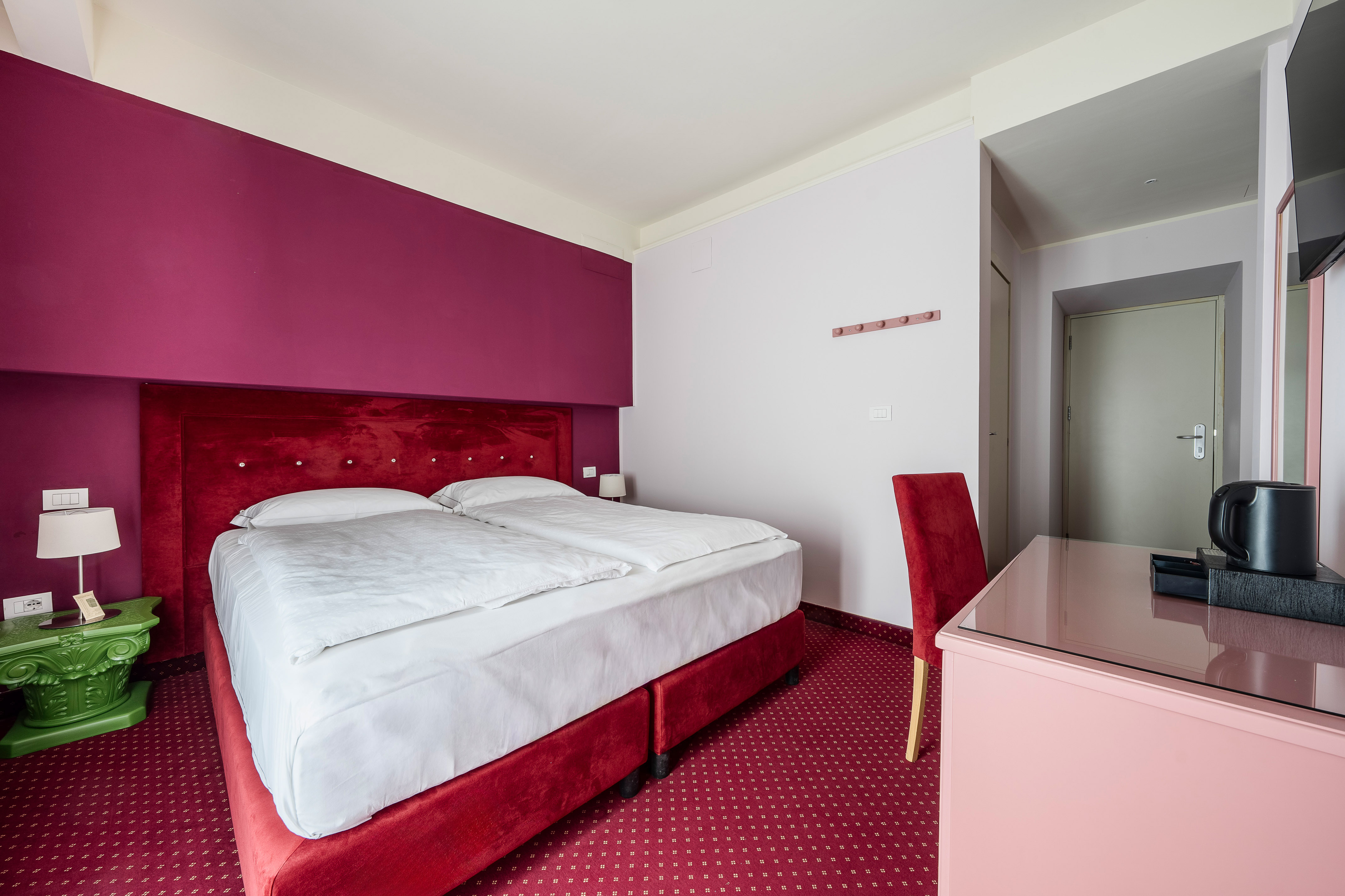 Hotel room with red velvet bed, magenta wall, and patterned red carpet.