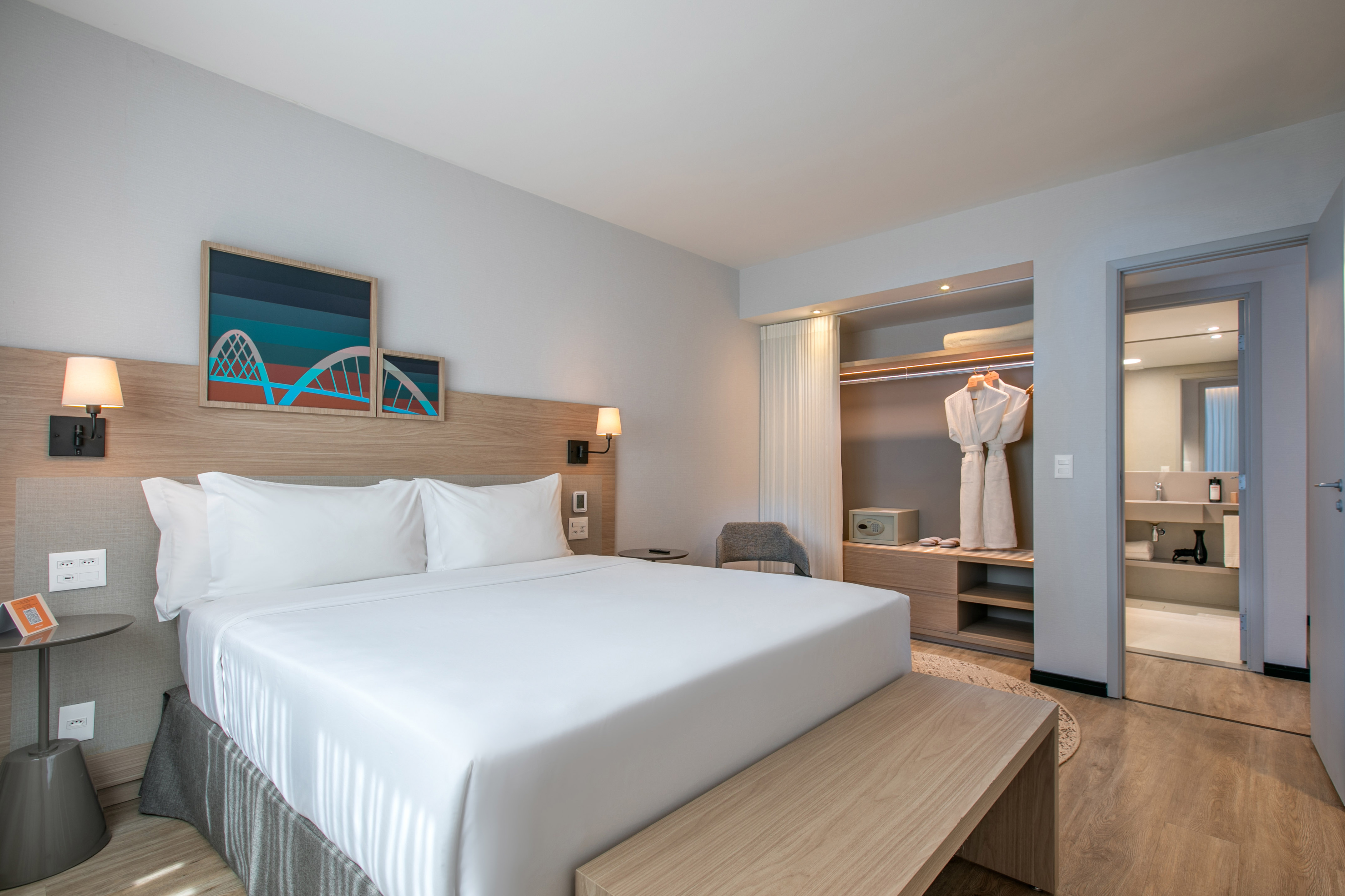 Modern hotel bedroom with white bed, wooden headboard, abstract art, and open closet.