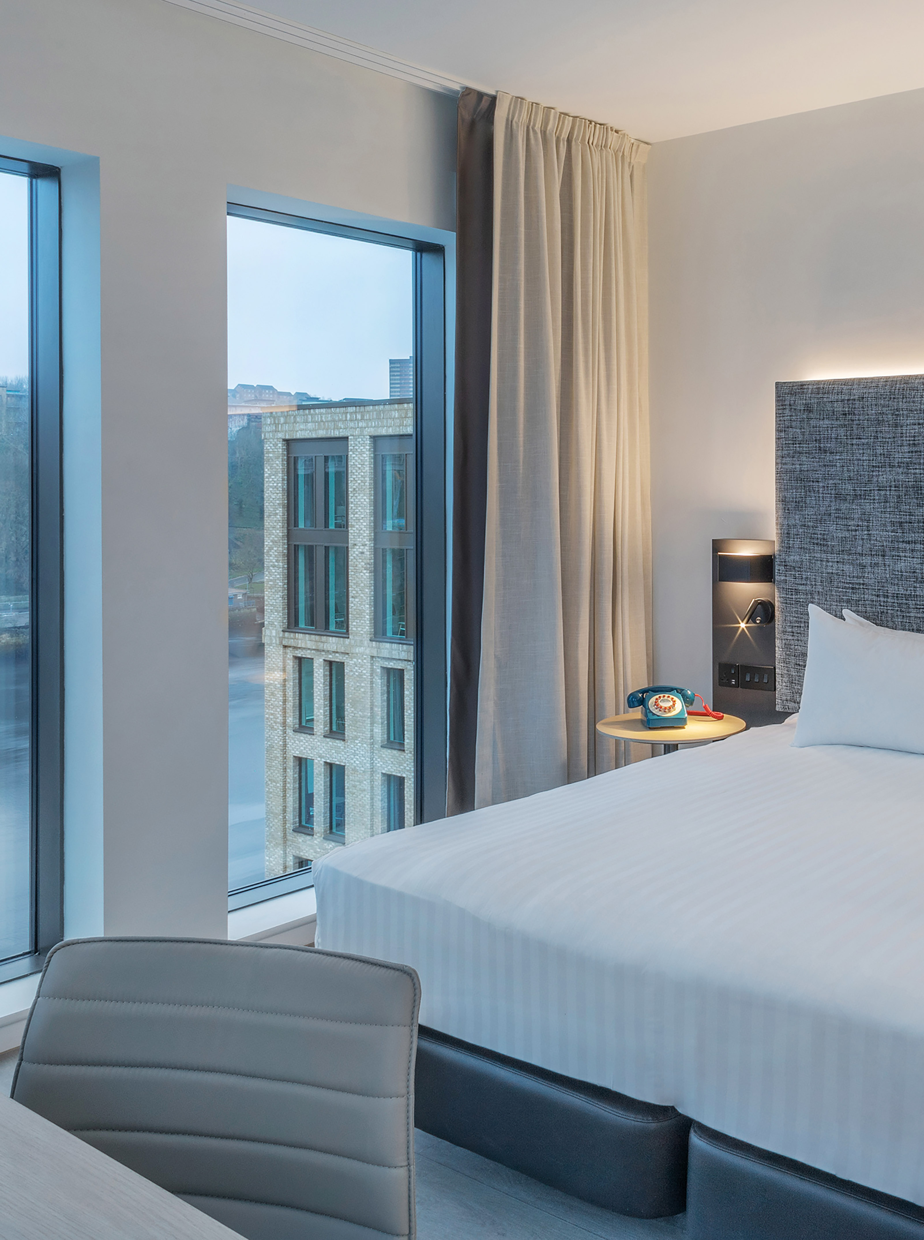 Modern hotel room, city view from large windows, white bed, and retro telephone.