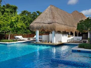 Vibrant resort pool with thatched swim-up bar, green trees.