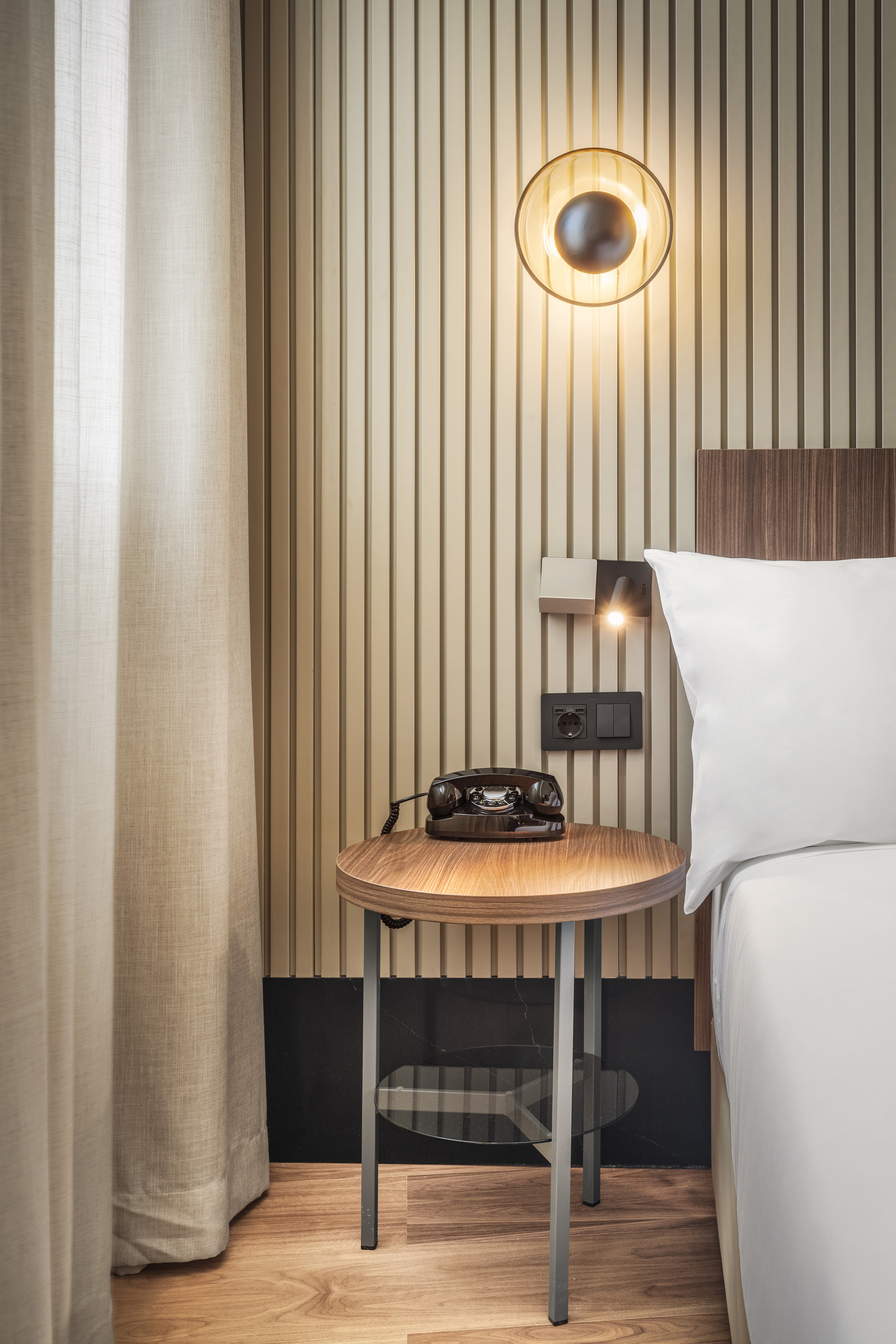 Hotel room bedside with vintage rotary telephone on round wood table.