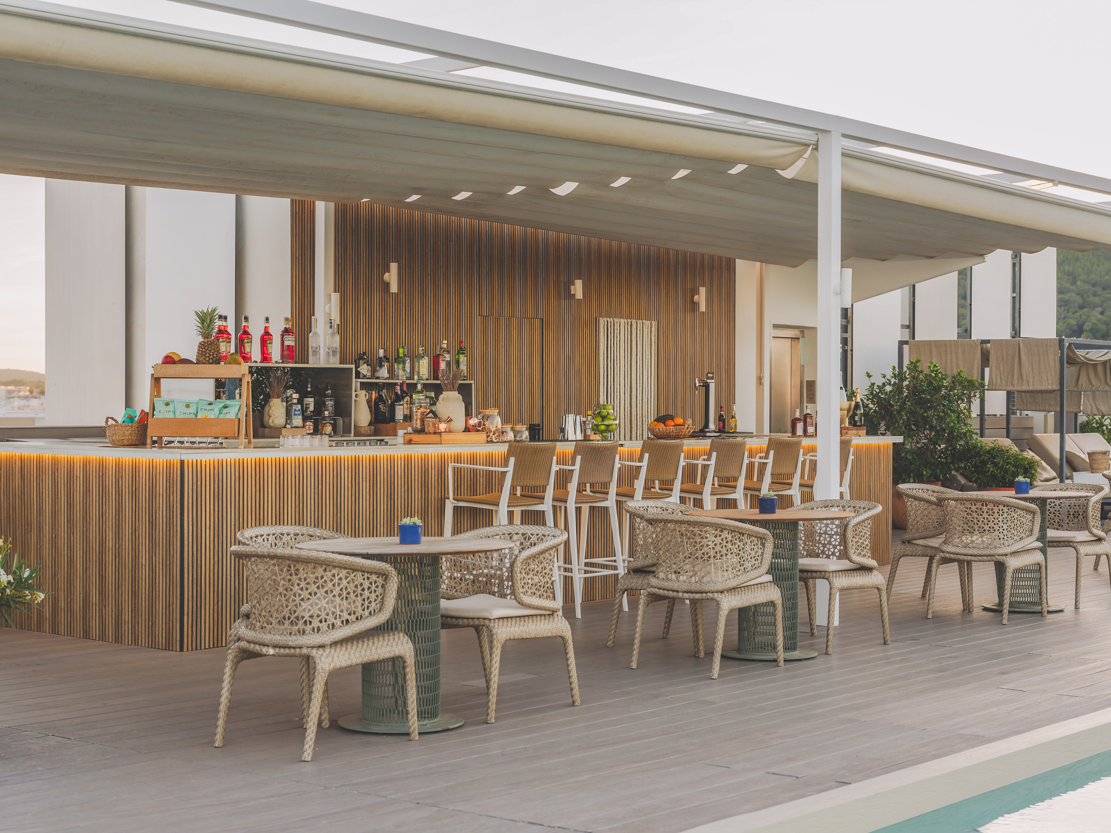 Modern outdoor hotel bar by pool with wooden counter and woven chairs.