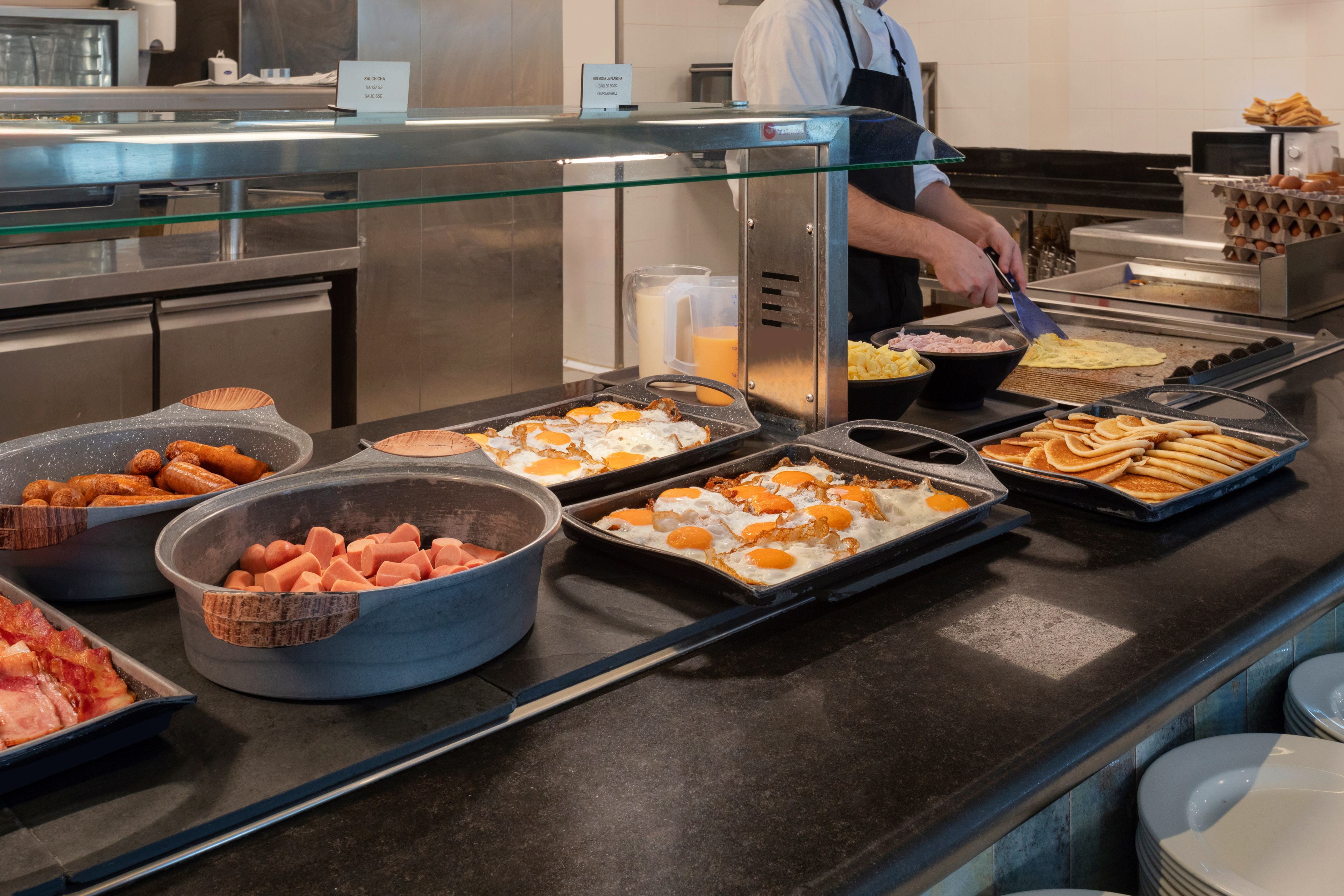 Chef cooking eggs at breakfast buffet with sausages, bacon, and pancakes.