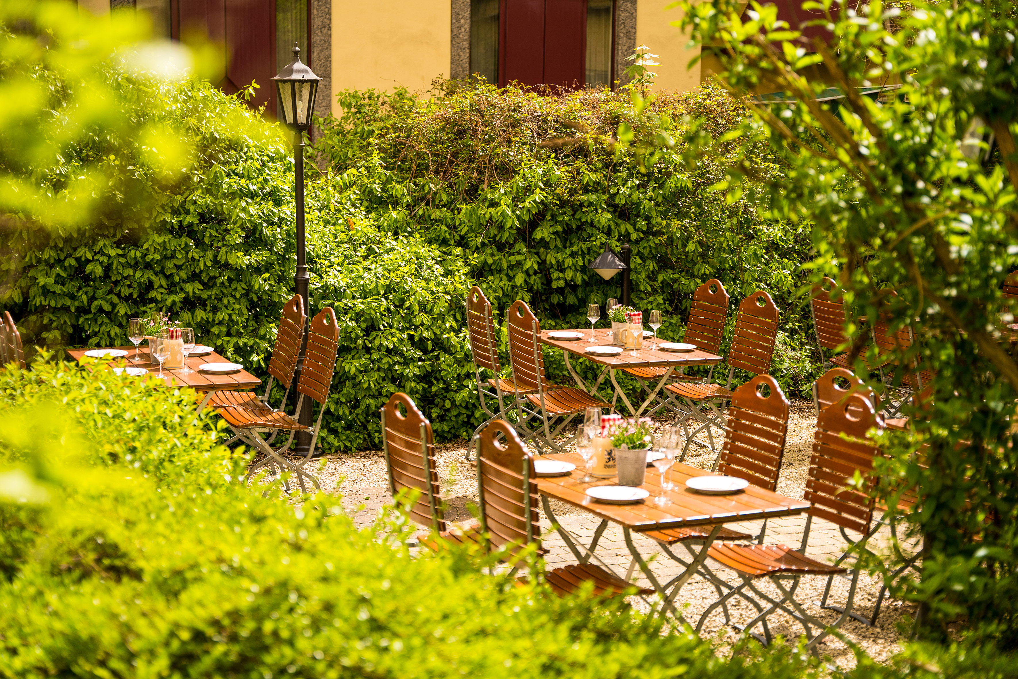 Outdoor dining area with wooden tables, chairs, and lush green hedges.