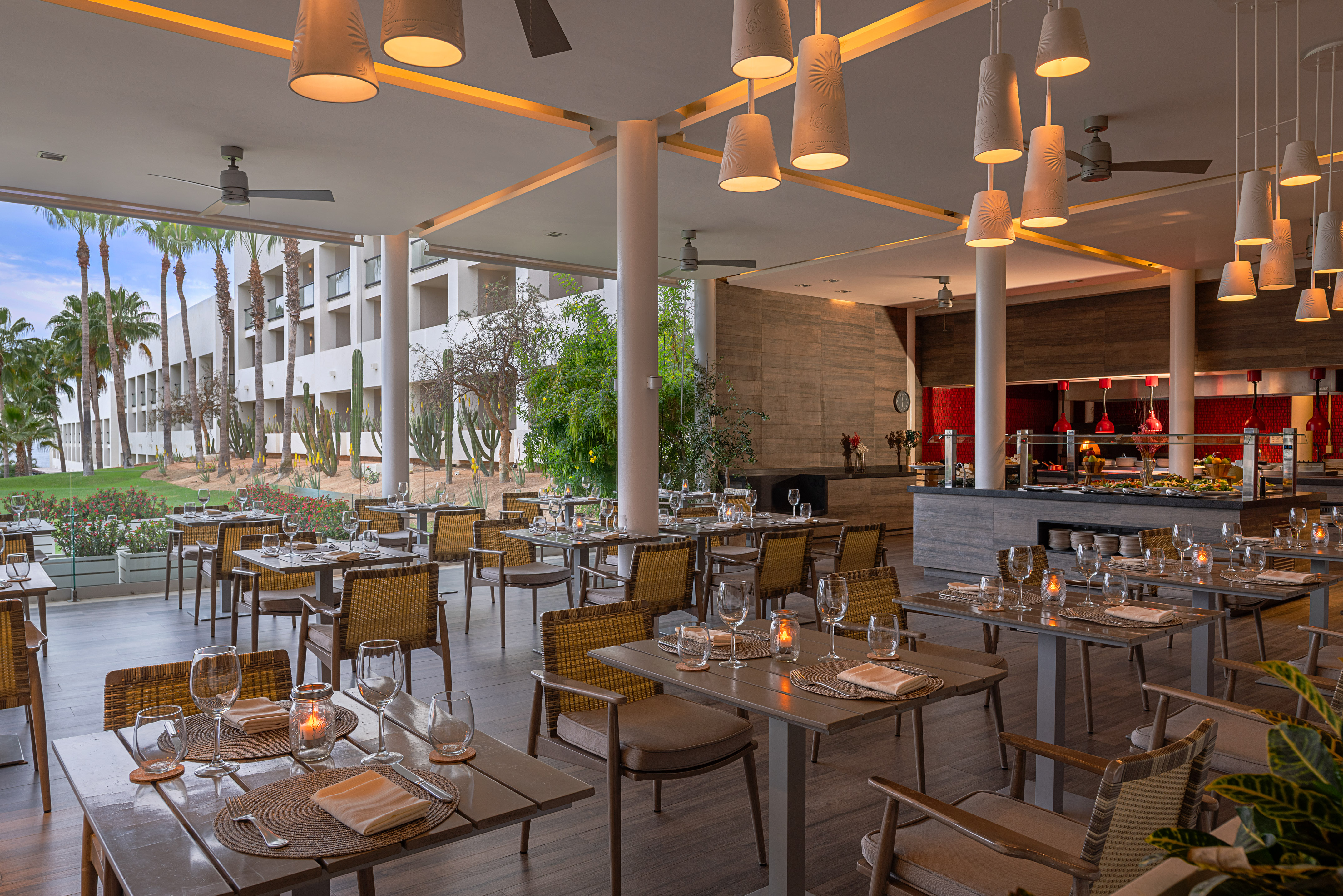 Modern resort restaurant with buffet, set tables, palm trees, and cacti view.