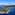 Helicopter flying above a green coastal island with yachts in a blue ocean.