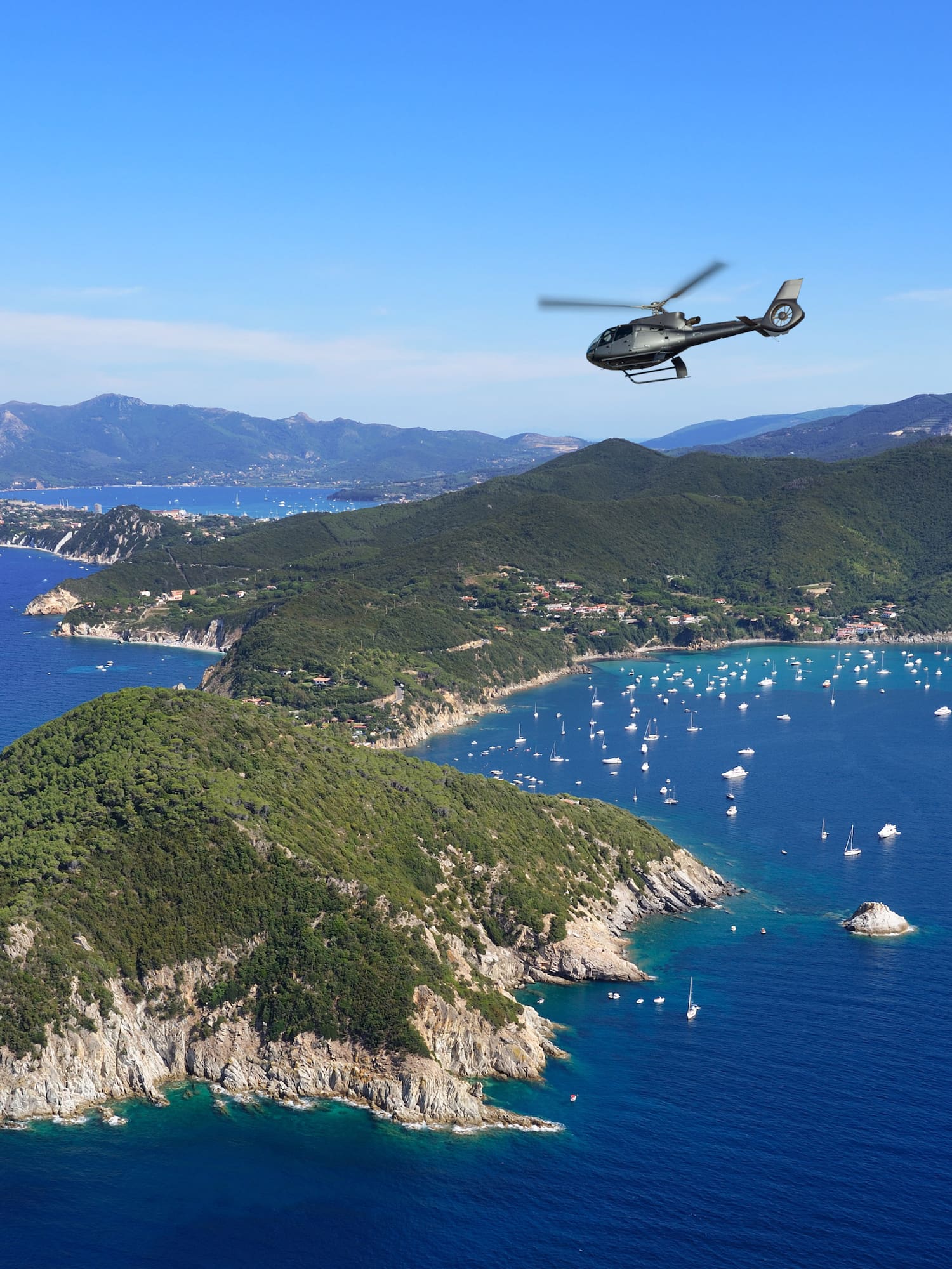 Helicopter flying above a green coastal island with yachts in a blue ocean.
