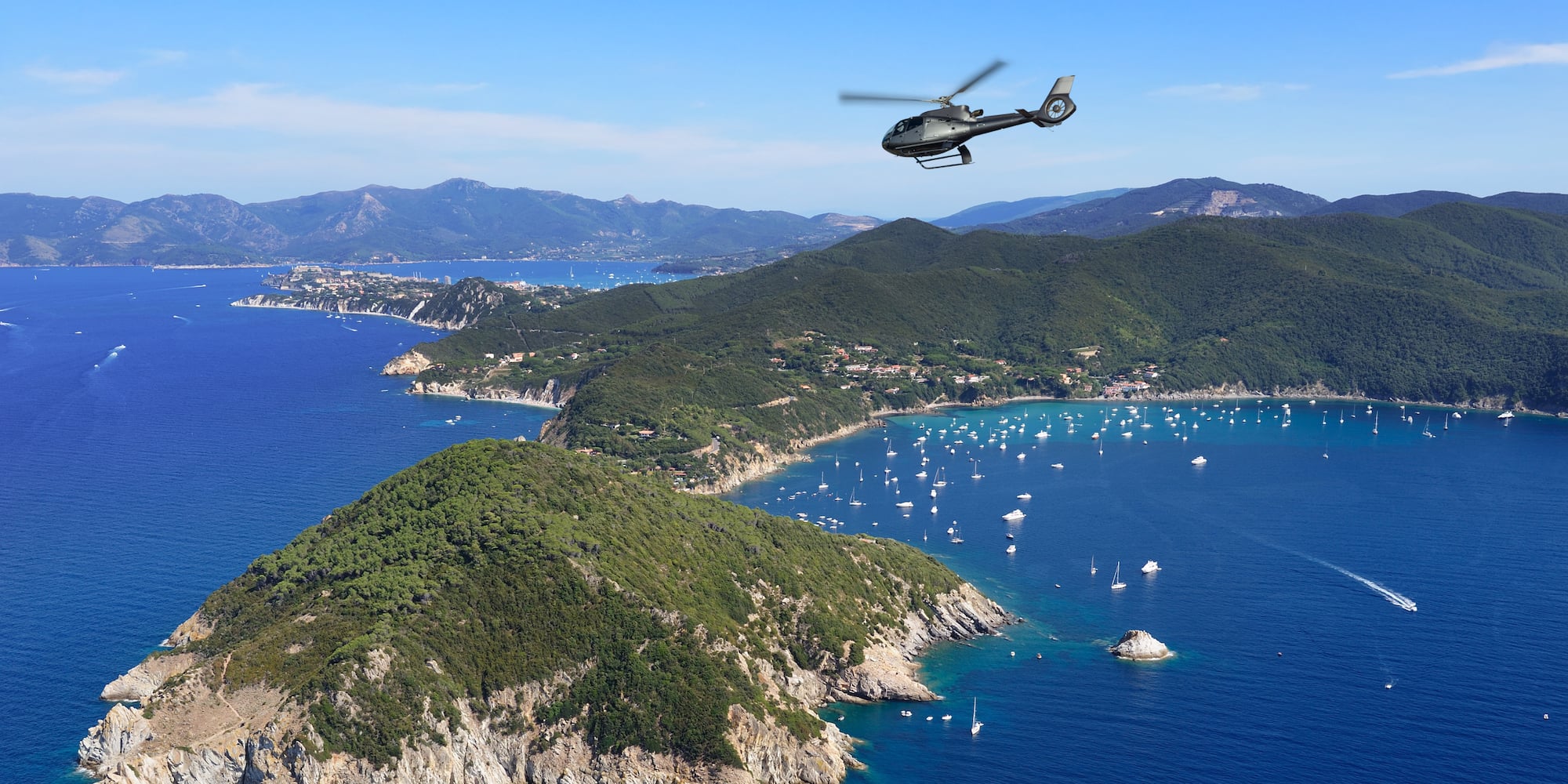 Helicopter flying above a green coastal island with yachts in a blue ocean.