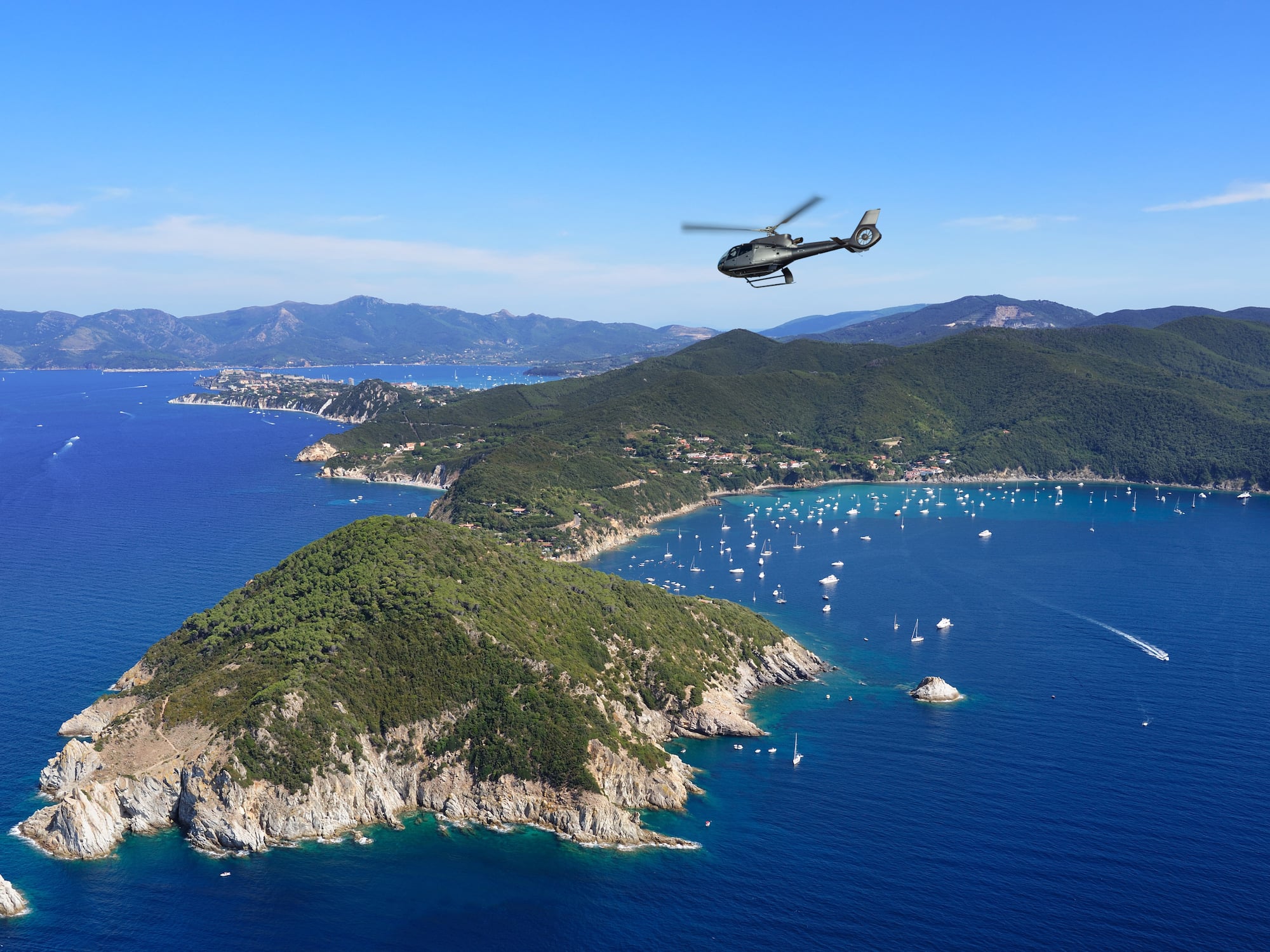 Helicopter flying above a green coastal island with yachts in a blue ocean.
