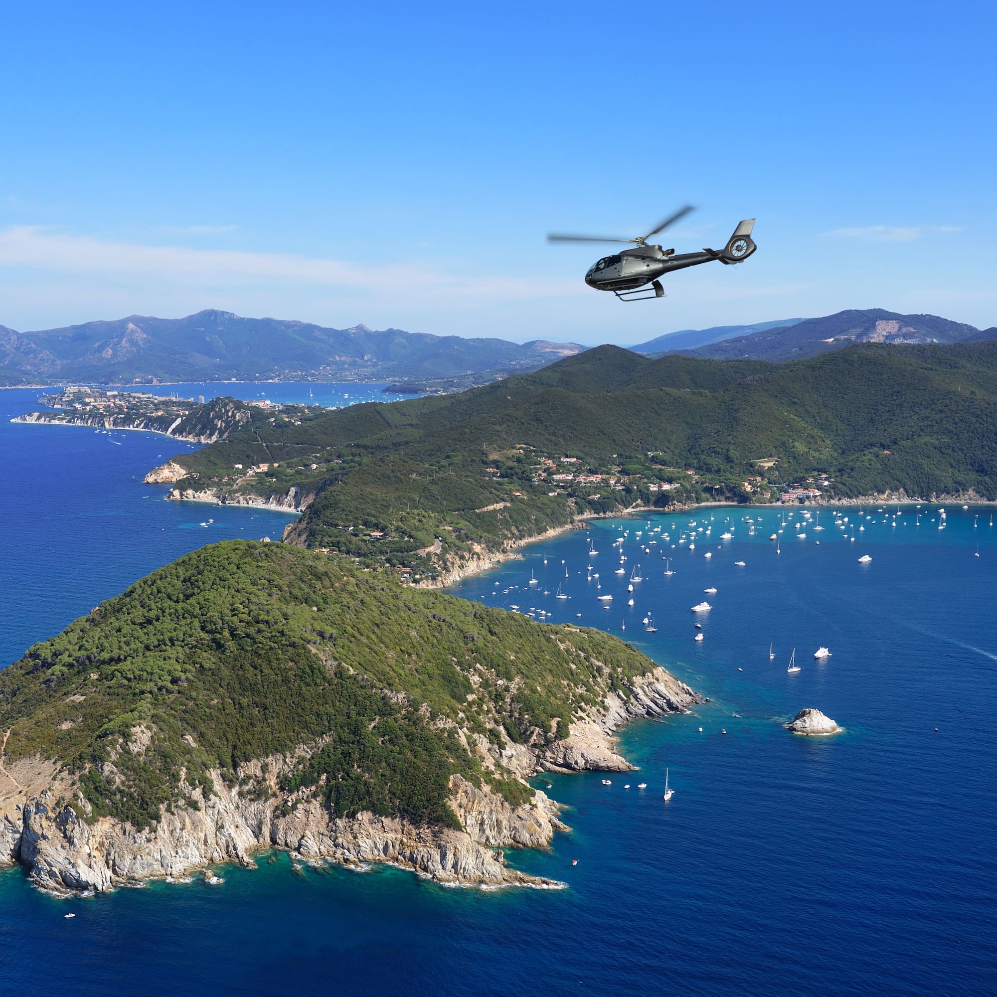 Helicopter flying above a green coastal island with yachts in a blue ocean.