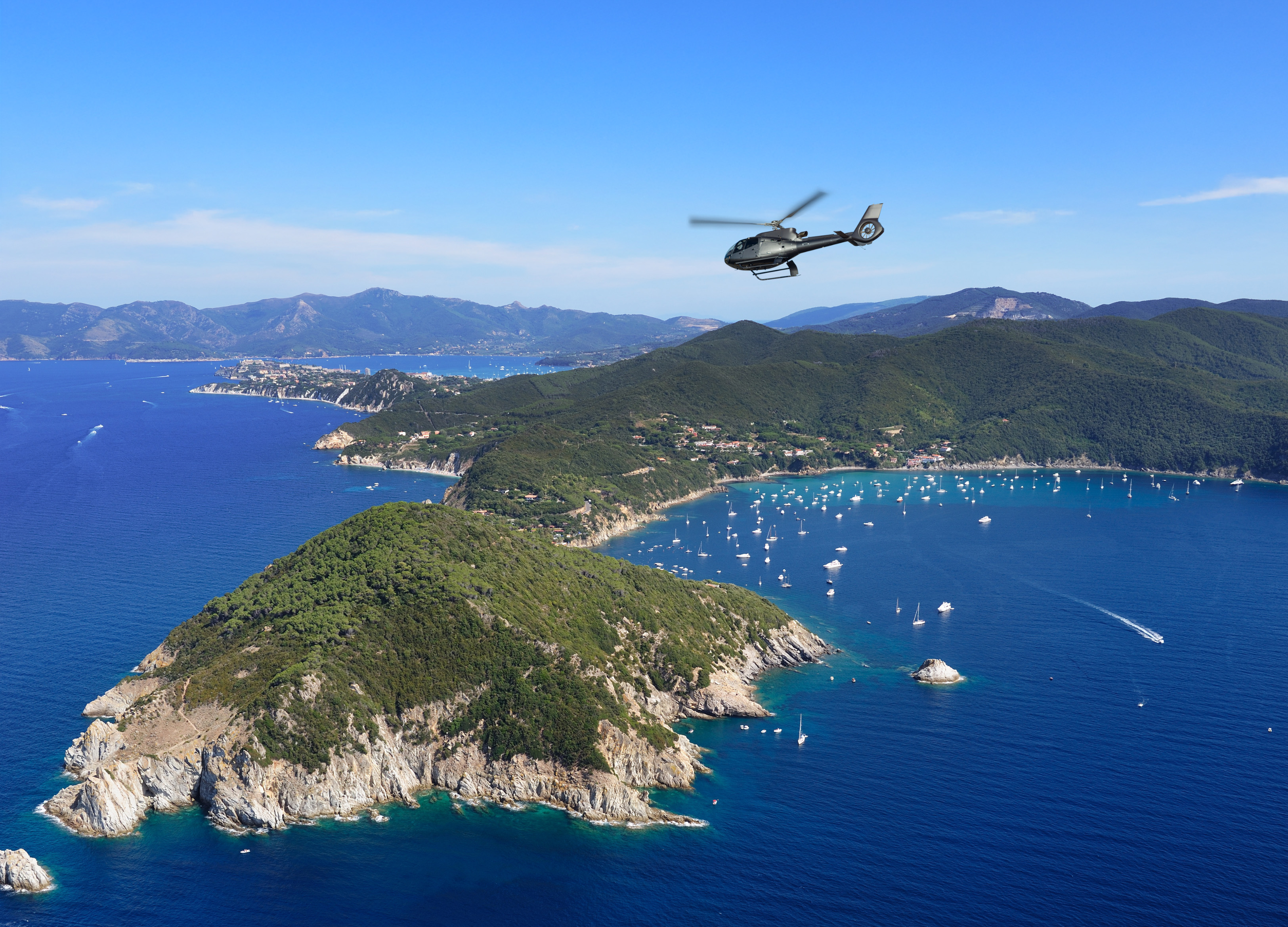 Helicopter flying above a green coastal island with yachts in a blue ocean.