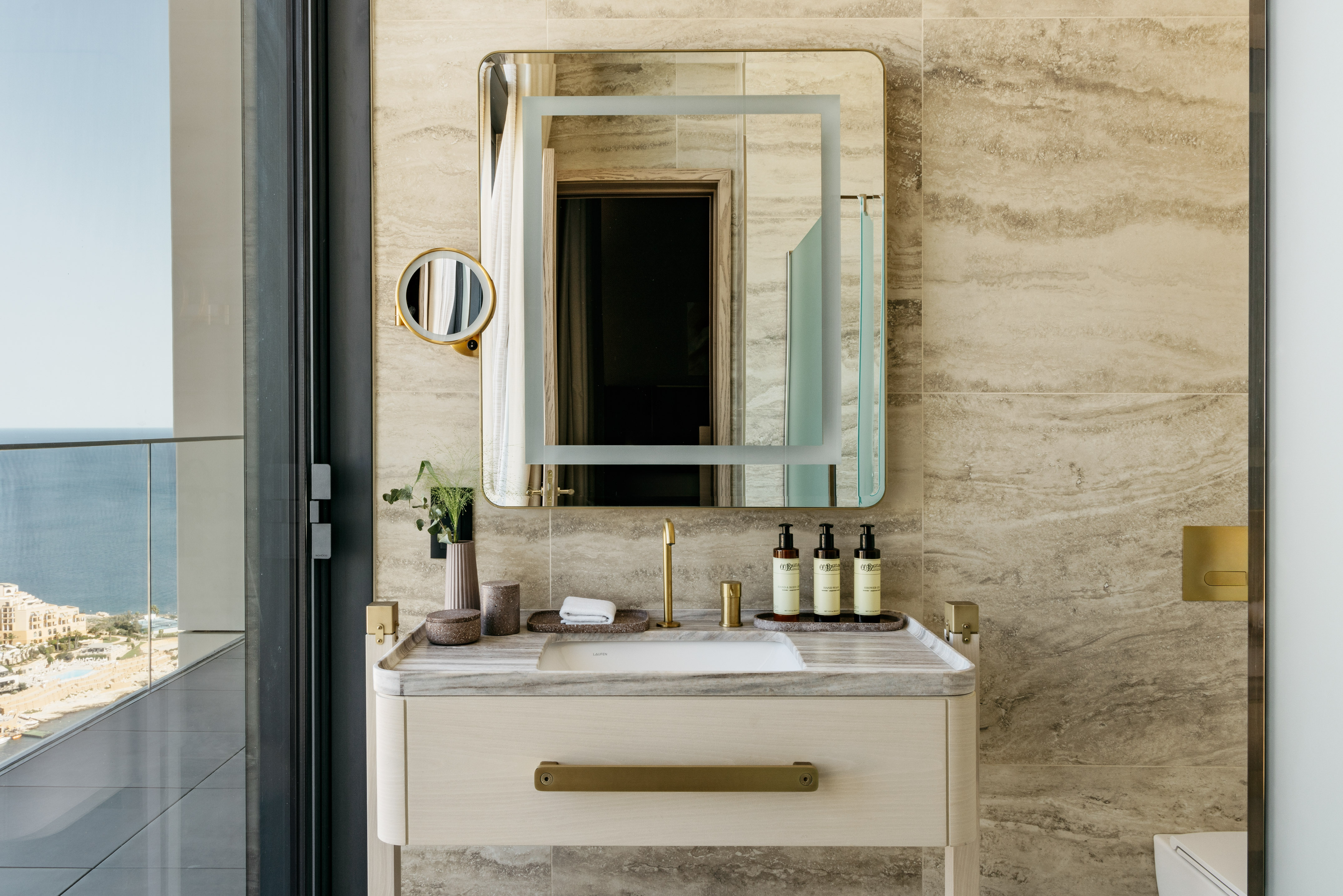 Luxury hotel bathroom with marble vanity, gold fixtures, backlit mirror, ocean view.
