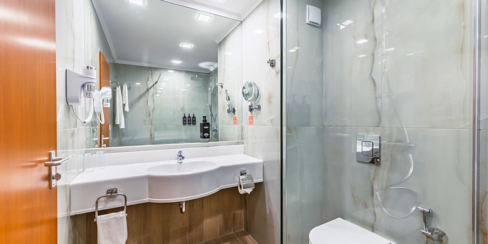 Modern hotel bathroom with white vanity, toilet, glass shower, and marble walls.