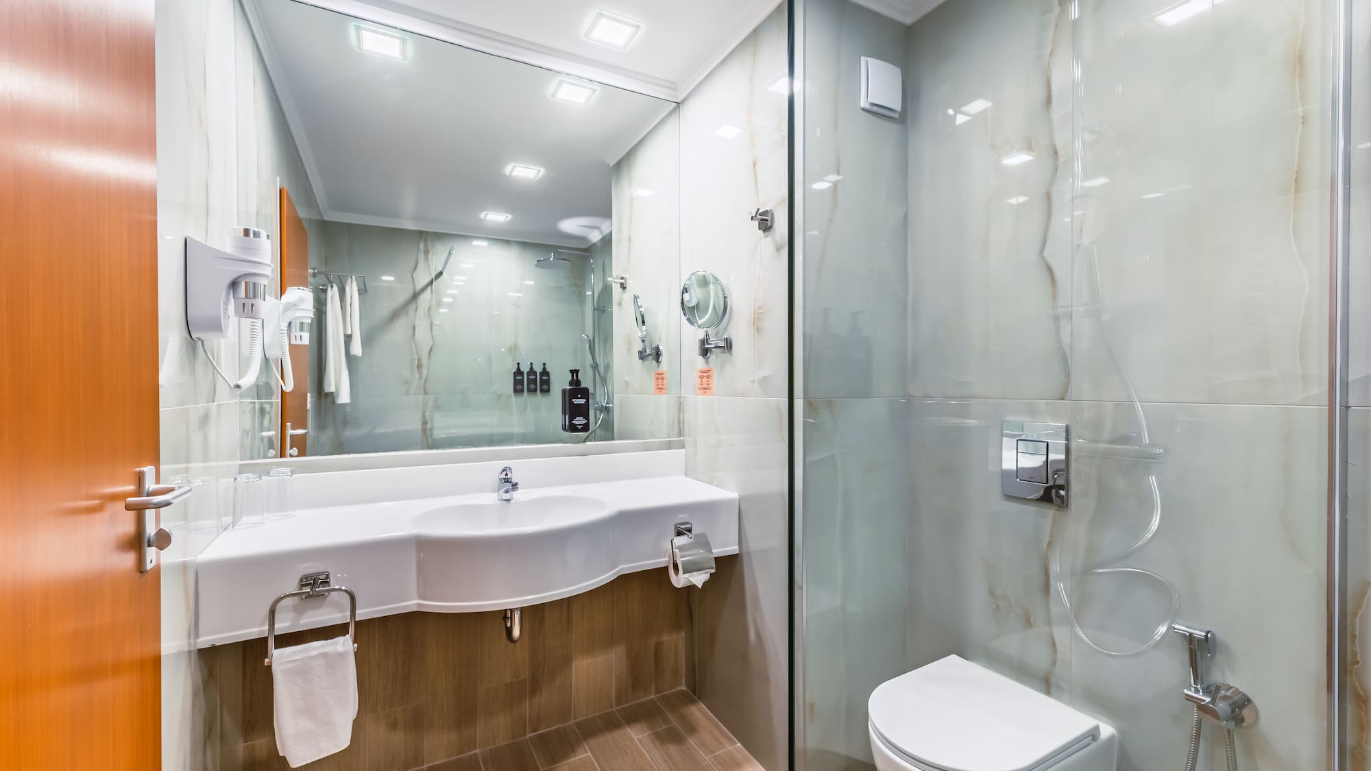 Modern hotel bathroom with white vanity, toilet, glass shower, and marble walls.