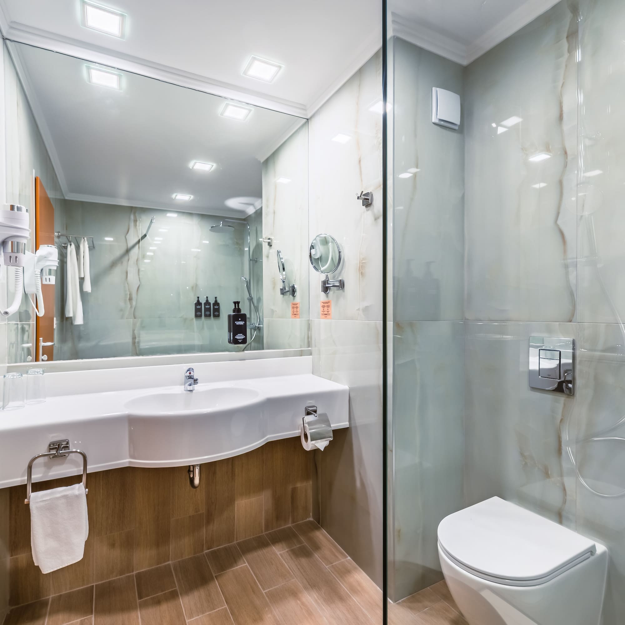 Modern hotel bathroom with white vanity, toilet, glass shower, and marble walls.