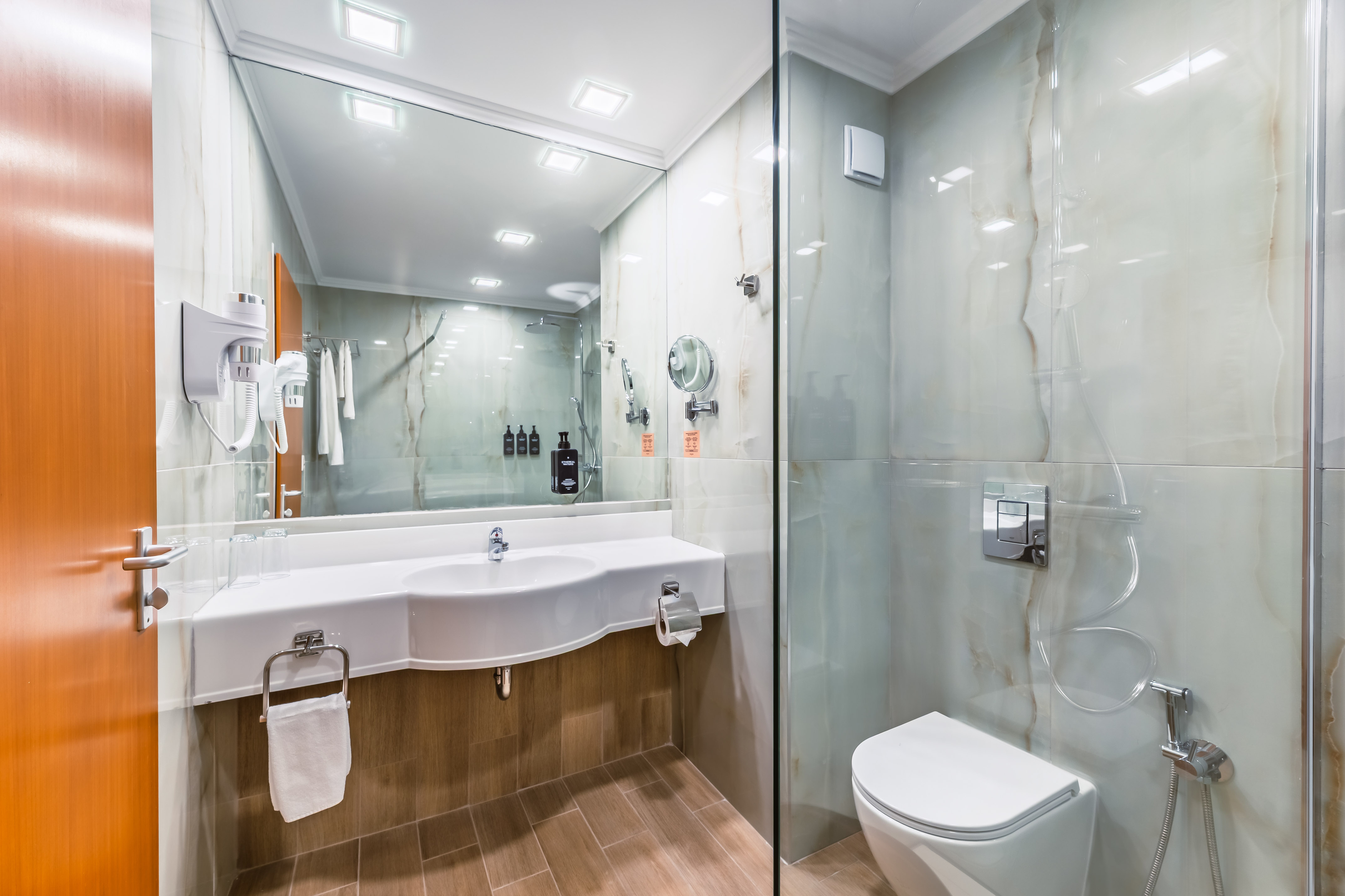 Modern hotel bathroom with white vanity, toilet, glass shower, and marble walls.