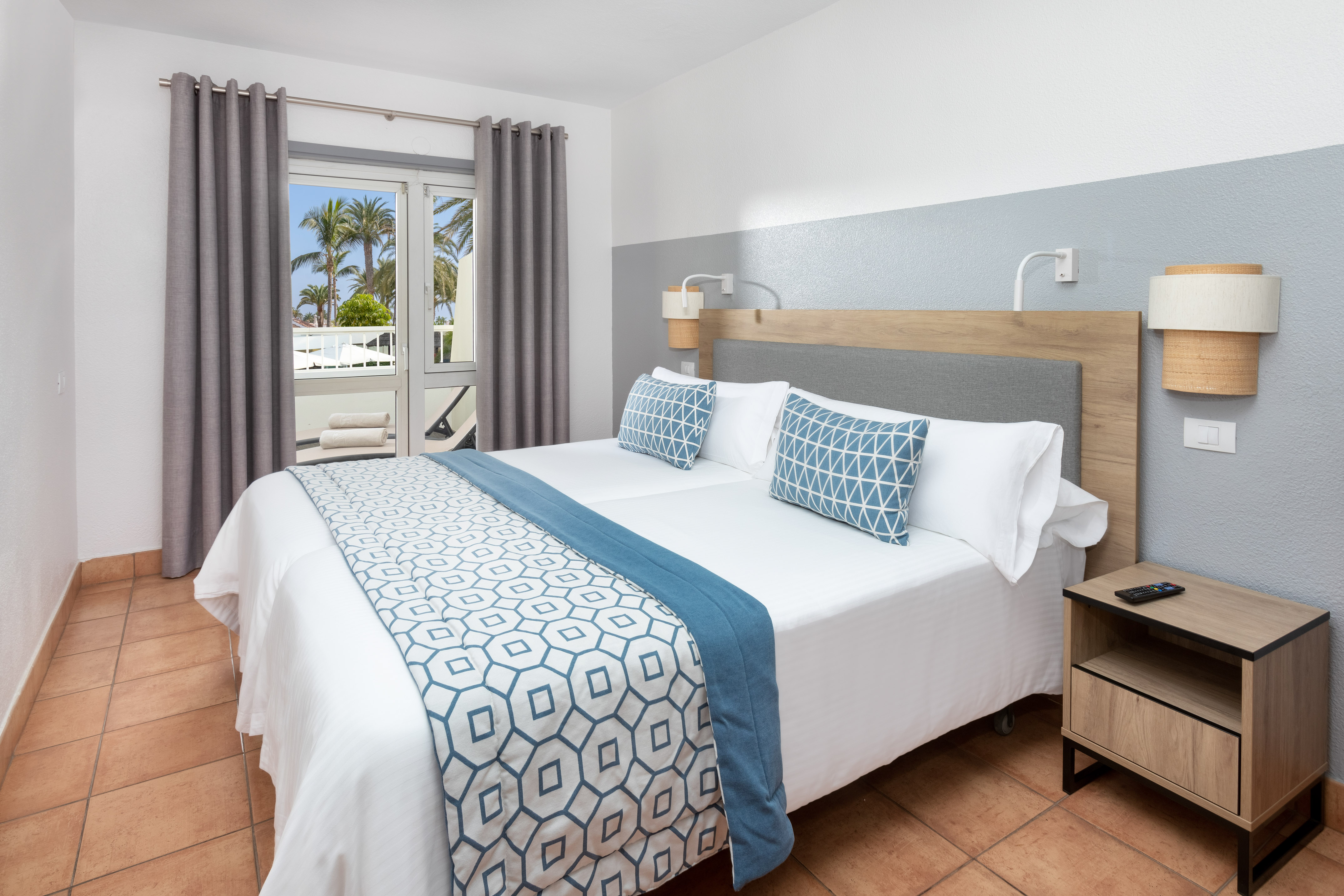Modern hotel room with twin beds, blue decor, and a palm tree view.