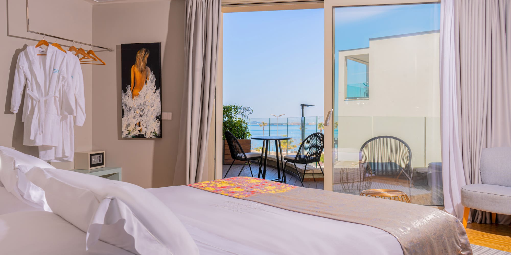 Bright hotel room with sea view balcony, white bed, and outdoor seating.
