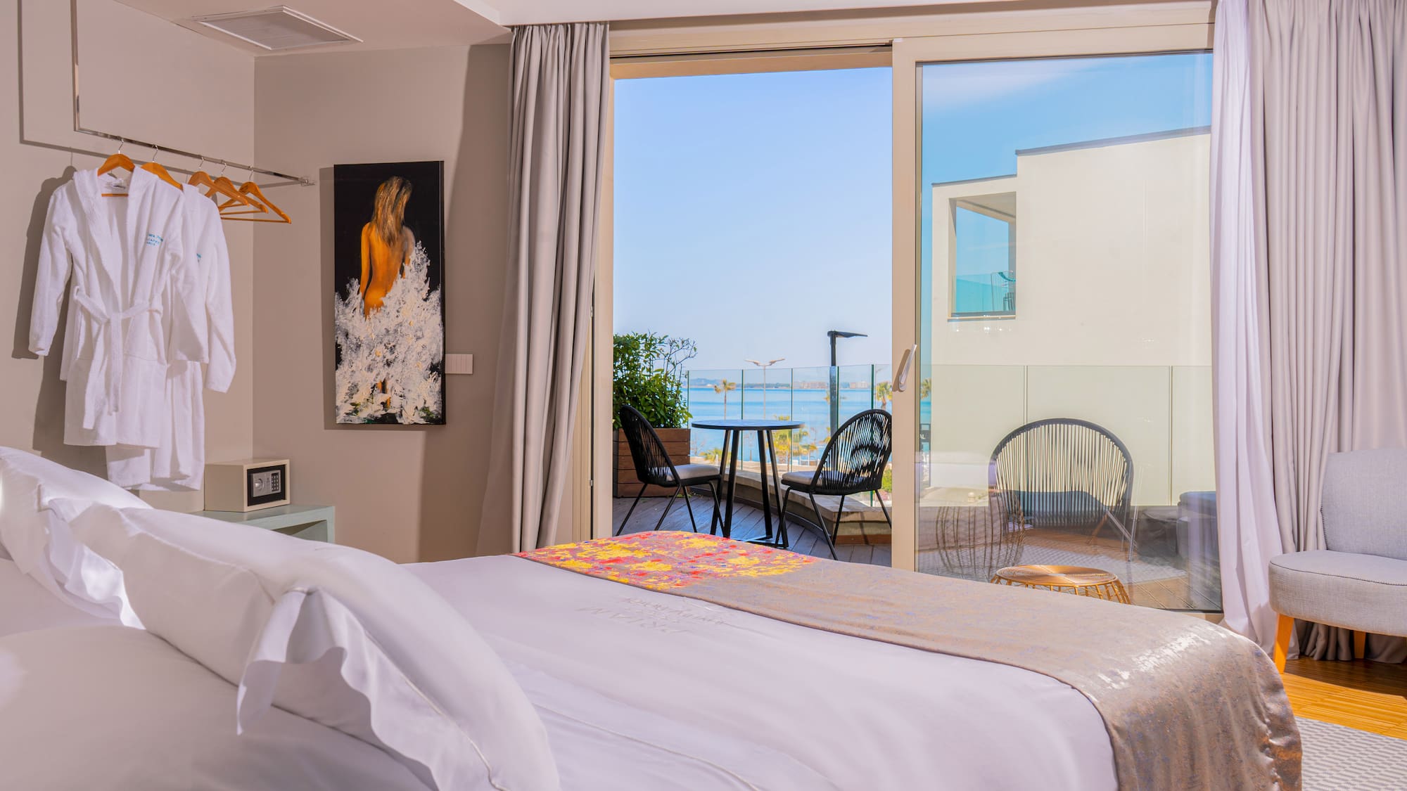 Bright hotel room with sea view balcony, white bed, and outdoor seating.