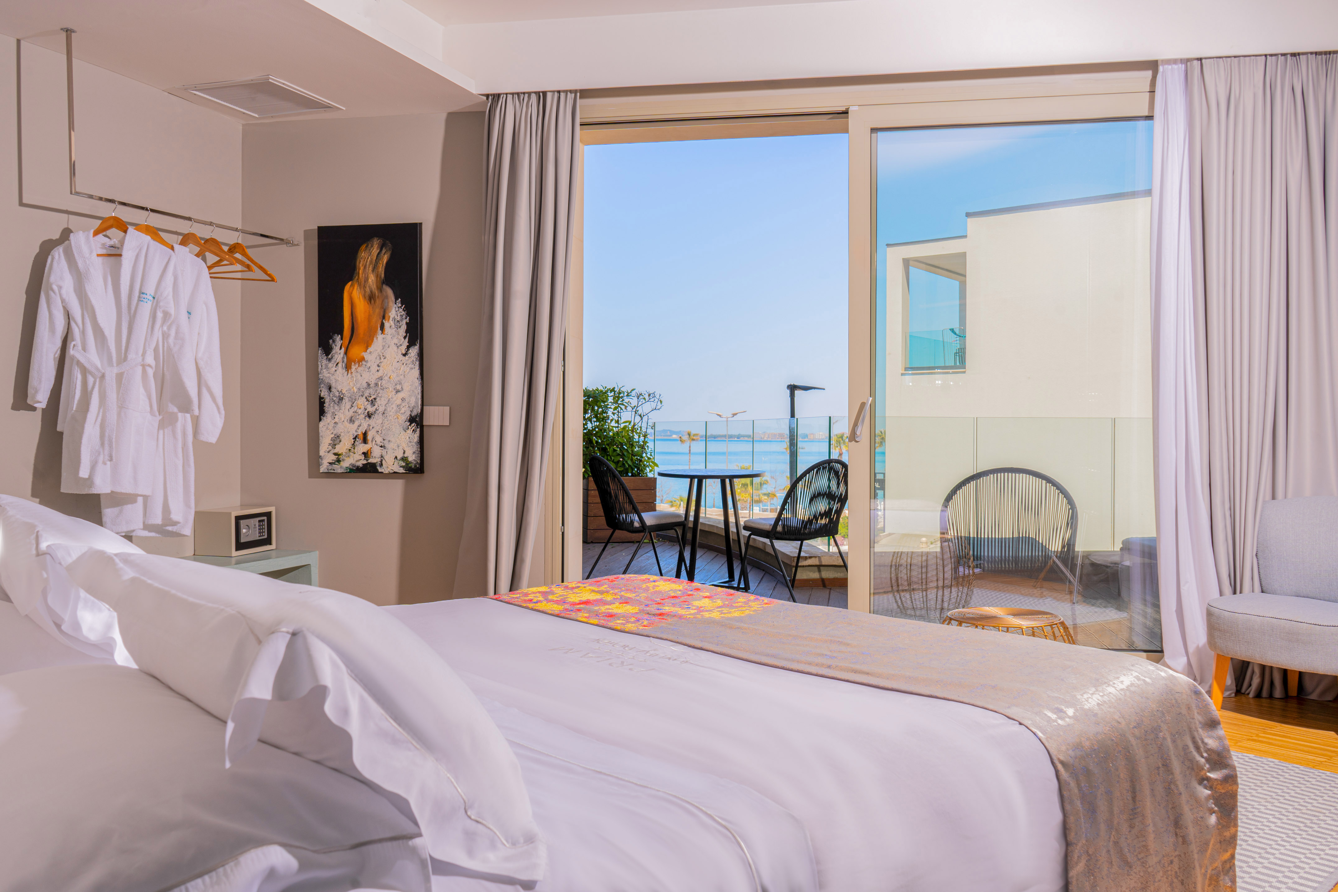 Bright hotel room with sea view balcony, white bed, and outdoor seating.