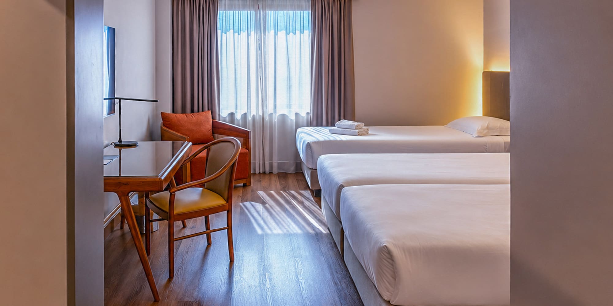 Bright triple hotel room with three single beds, desk, and armchair.