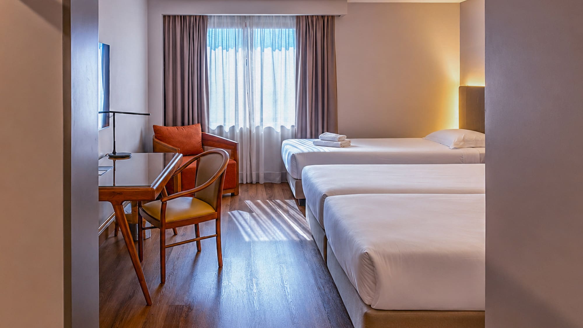 Bright triple hotel room with three single beds, desk, and armchair.