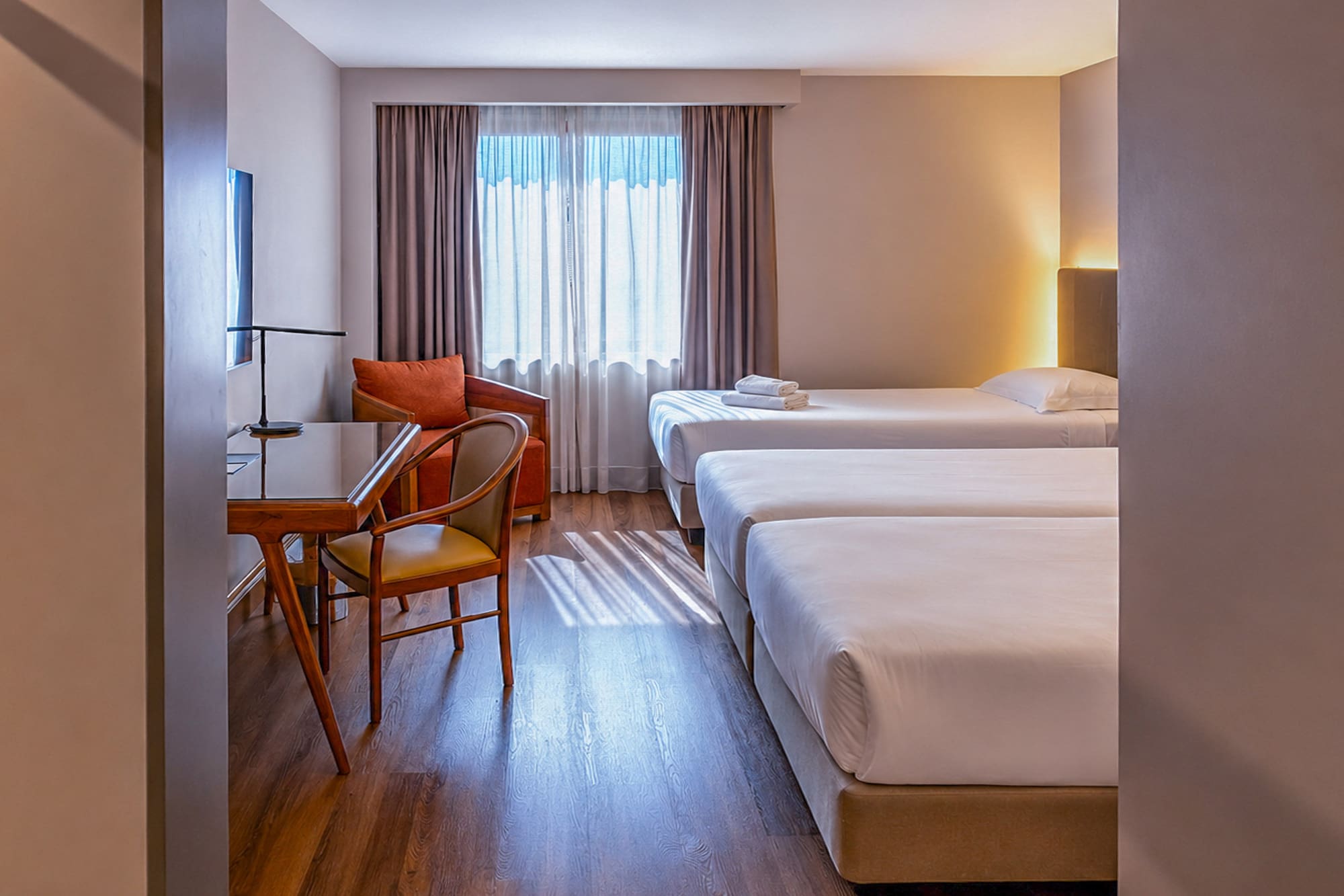 Bright triple hotel room with three single beds, desk, and armchair.