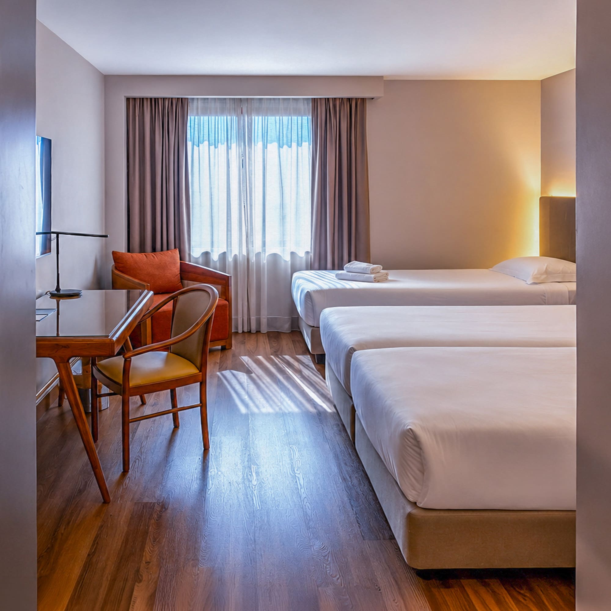 Bright triple hotel room with three single beds, desk, and armchair.