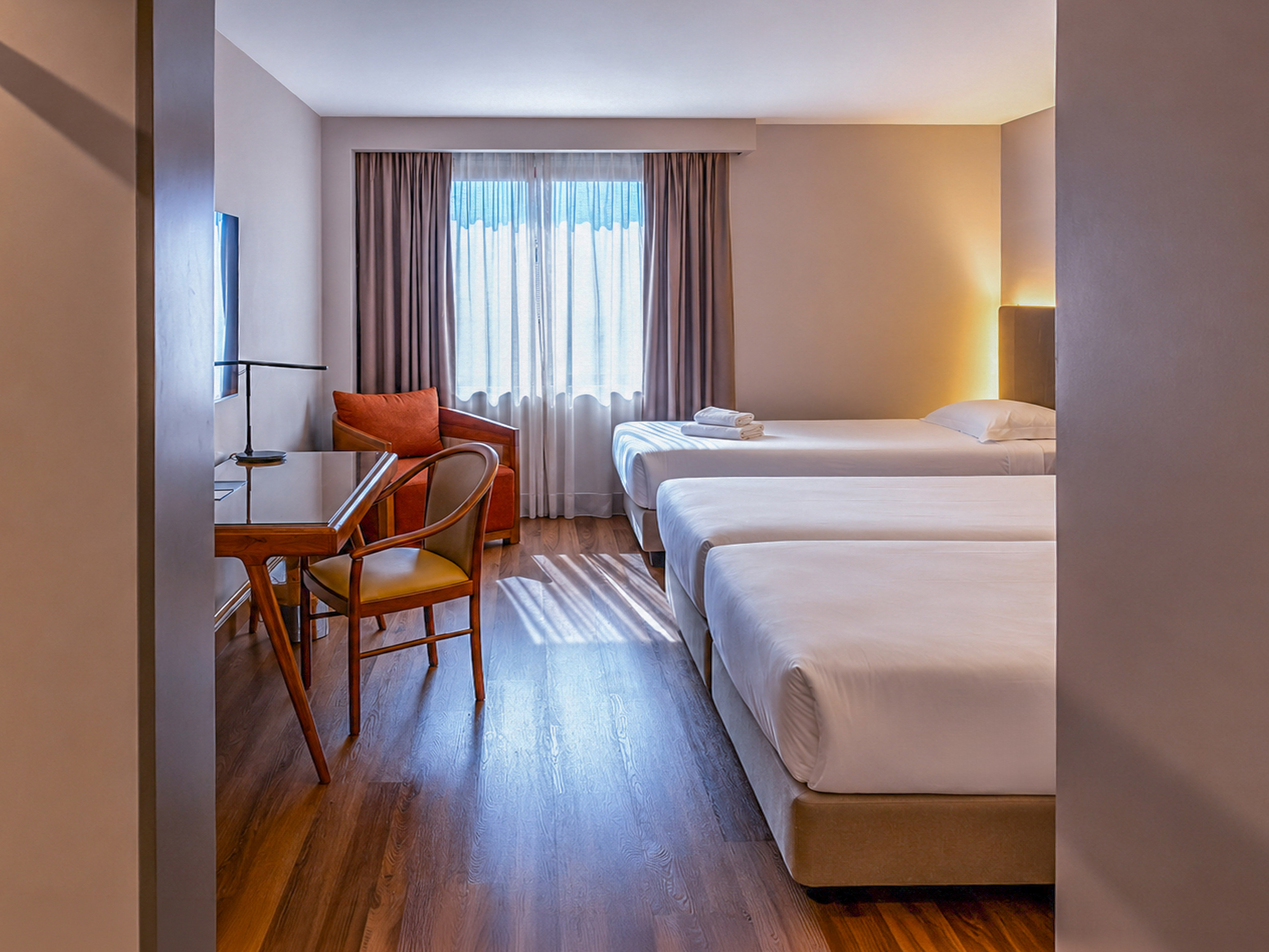 Bright triple hotel room with three single beds, desk, and armchair.
