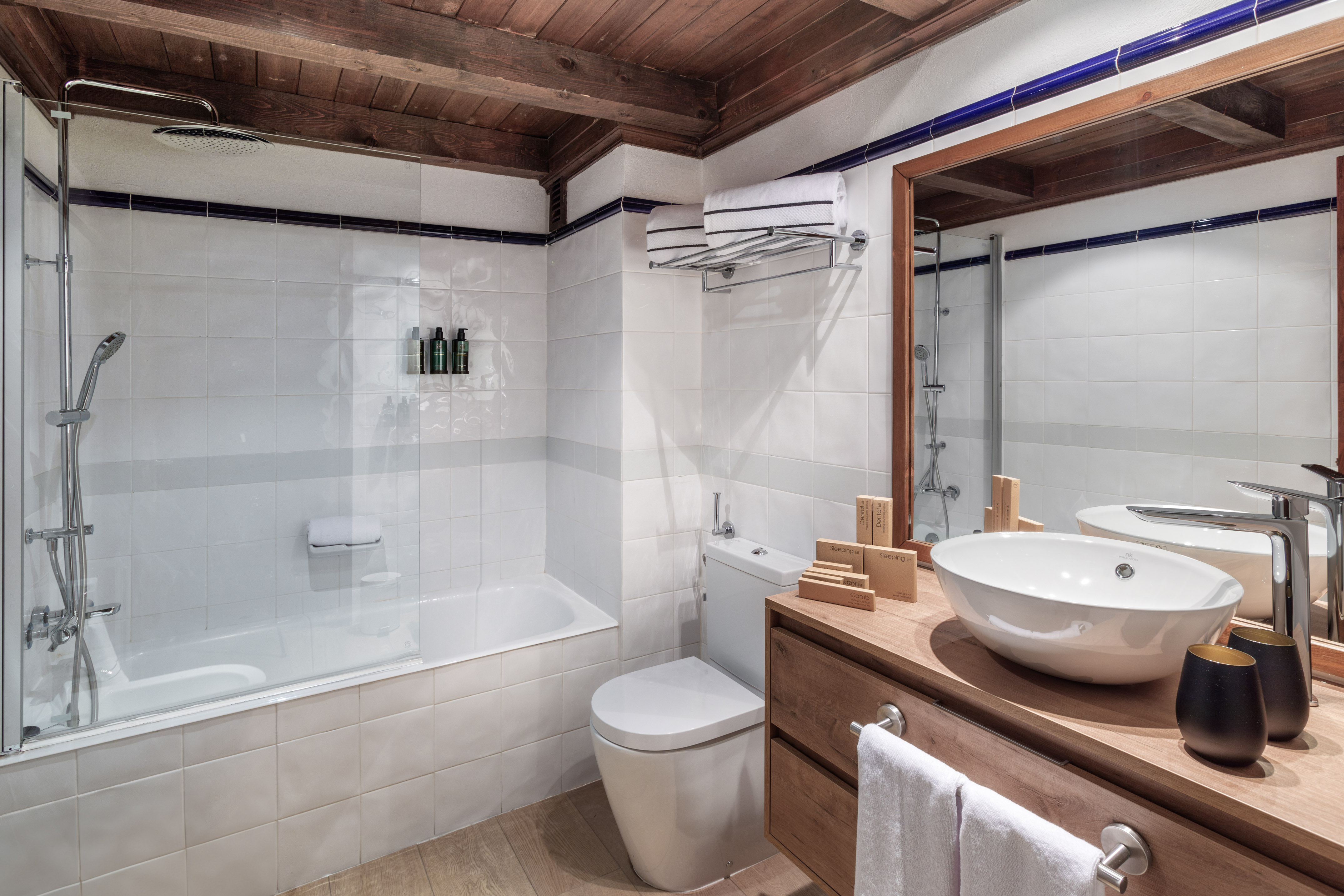 White tiled hotel bathroom with wooden ceiling, bathtub, and vessel sink.
