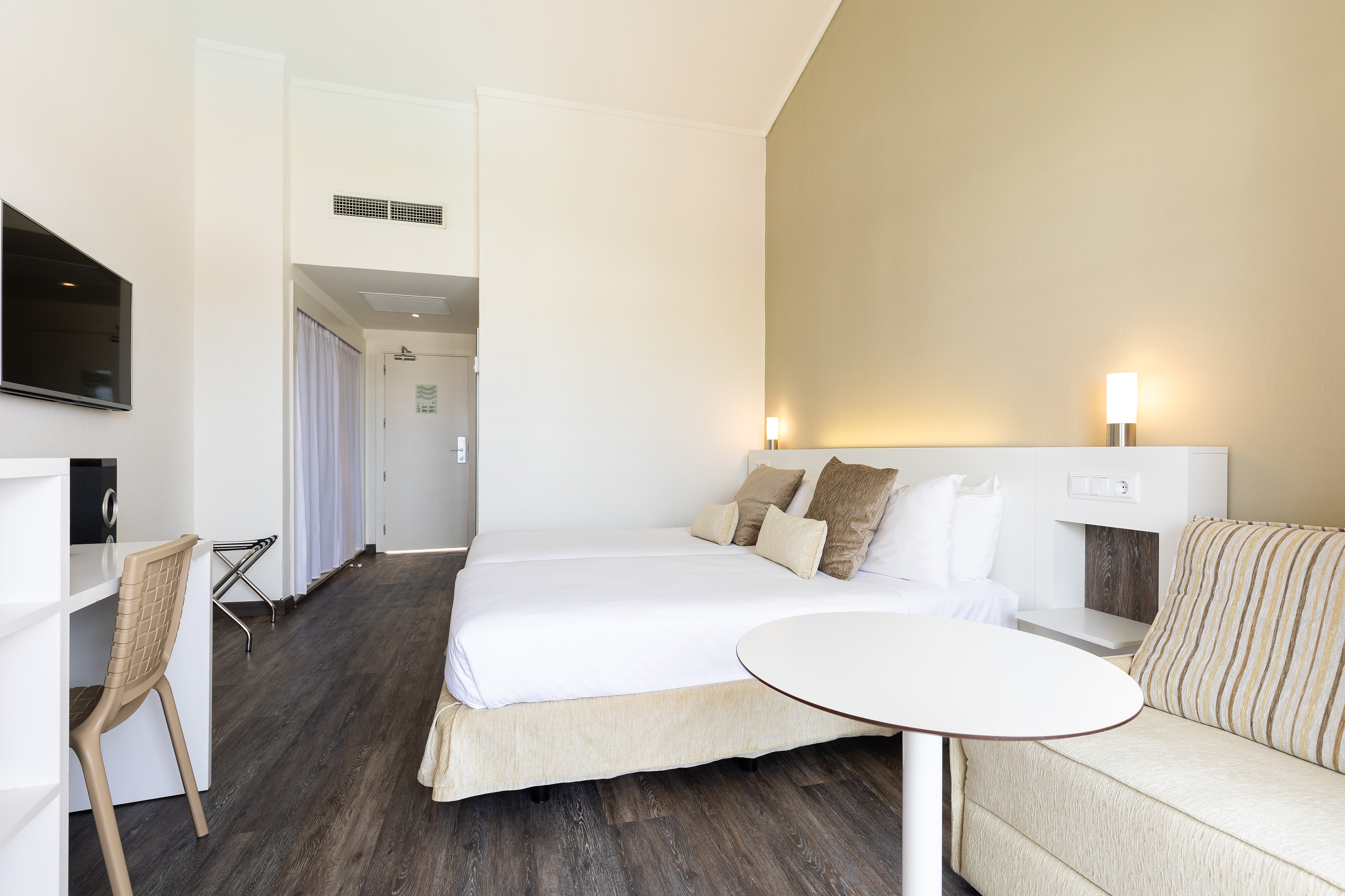 Modern hotel room with twin beds, desk, TV, and sofa.